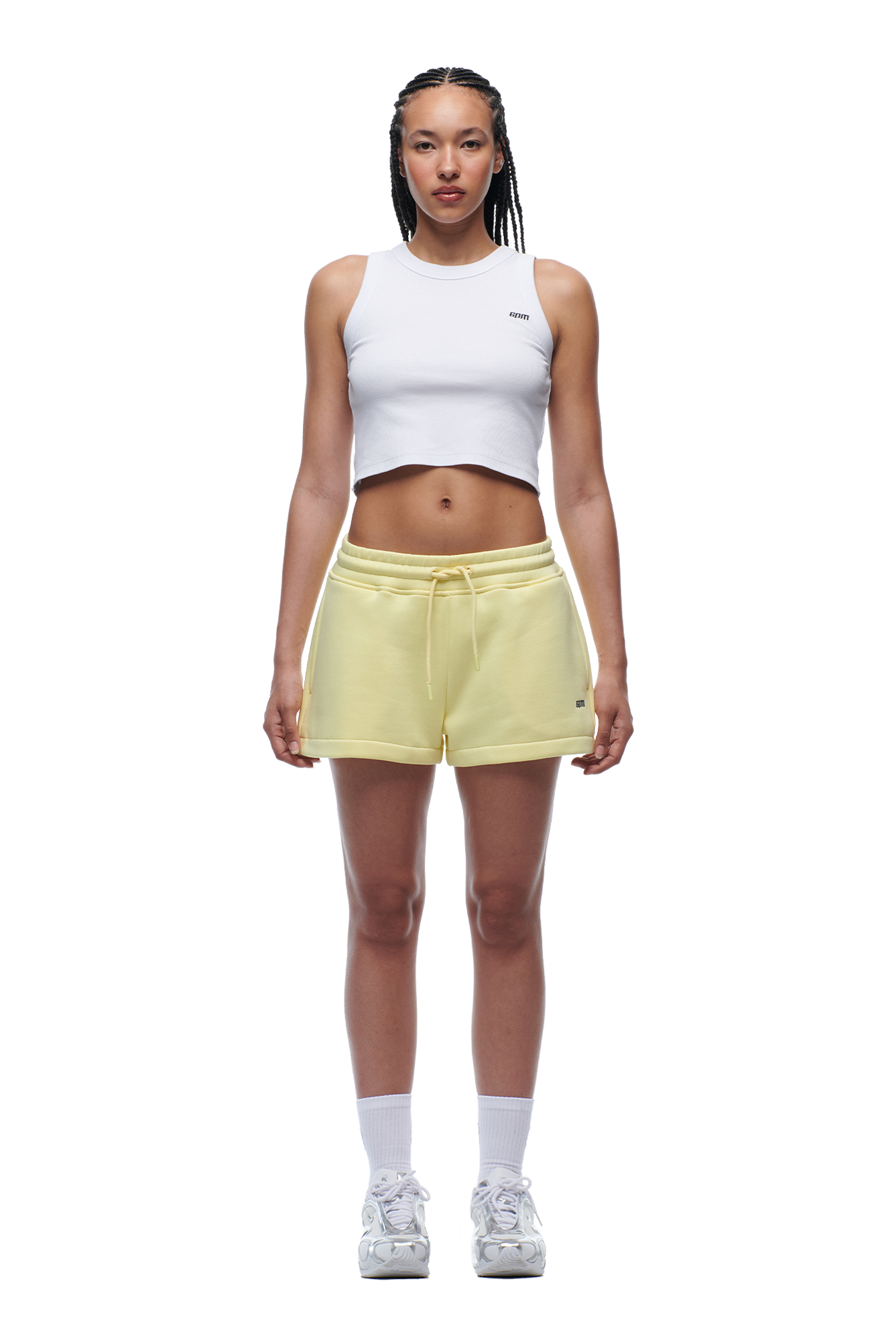 A woman with braided hair and a neutral expression faces forward, wearing a white cropped tank top, 6PM WMNS SHORTS BUTTERCREAM with white side stripes, white socks, and white sneakers.