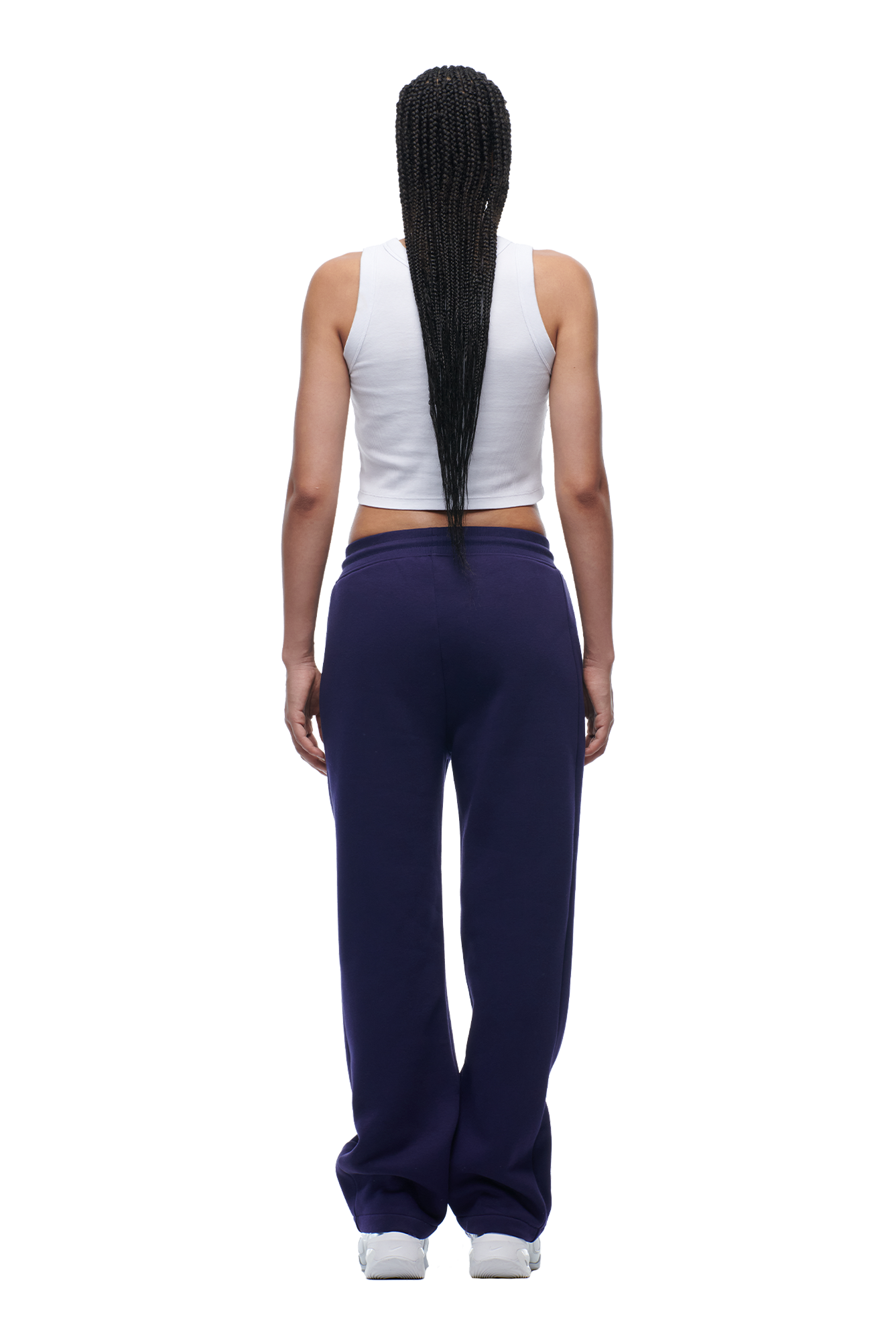 A person with long braided hair stands facing away, wearing a white sleeveless crop top, 6PM WMNS OPEN LEG ORBIT BLUE pants, and white sneakers.