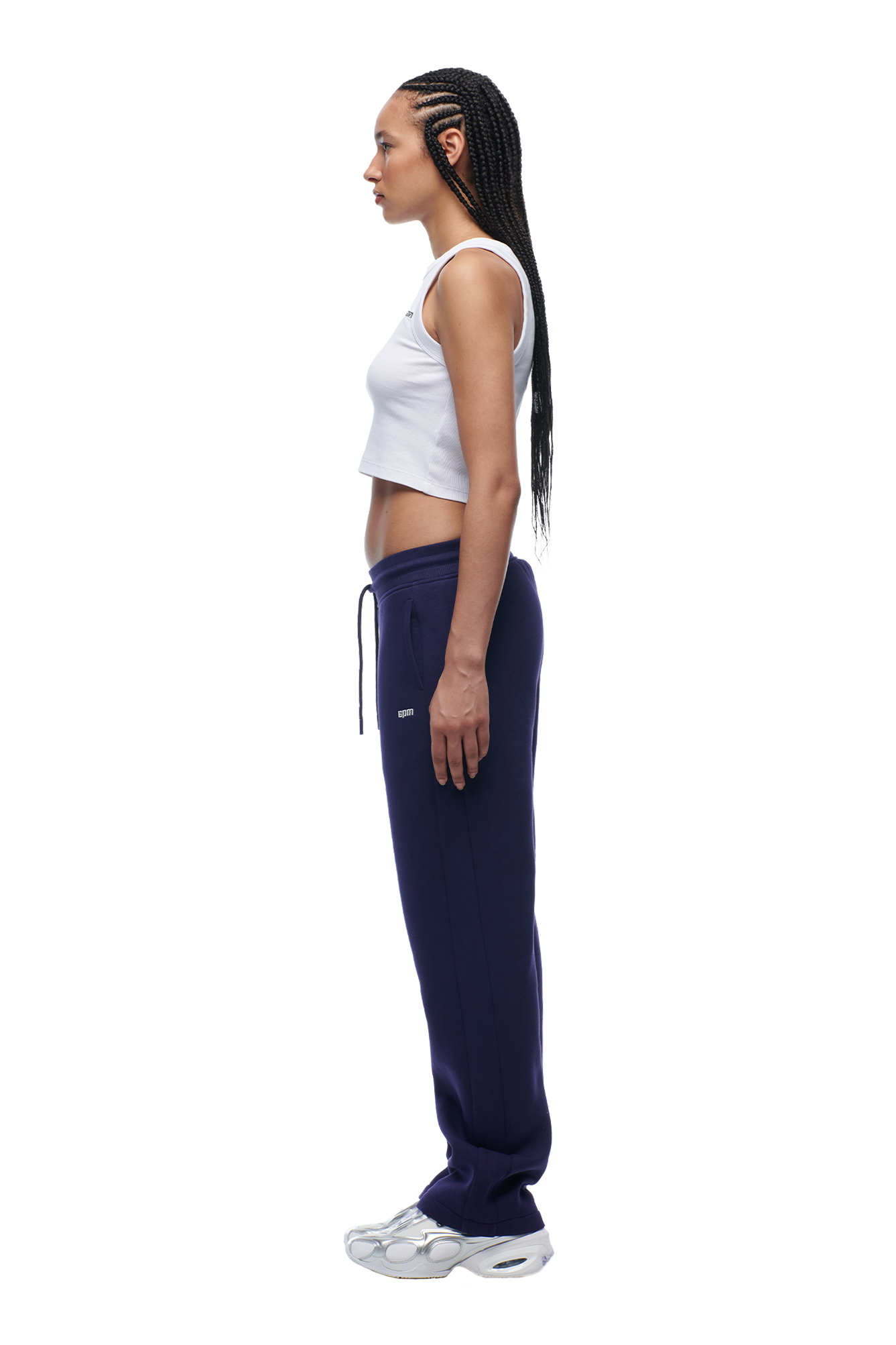 A woman with long braided hair stands in profile, wearing the 6PM WMNS OPEN LEG ORBIT BLUE sweatpants, a white crop top, and white sneakers against a white background.