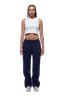 A woman with braided hair models the 6PM WMNS OPEN LEG ORBIT BLUE sweatpants, paired with a white crop top and white sneakers, standing forward-facing against a white background.