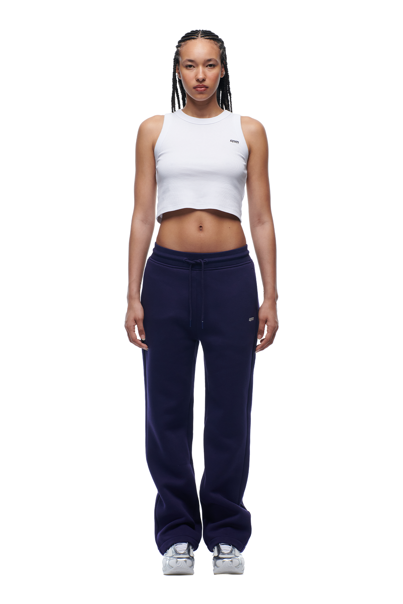 A woman with braided hair models the 6PM WMNS OPEN LEG ORBIT BLUE sweatpants, paired with a white crop top and white sneakers, standing forward-facing against a white background.