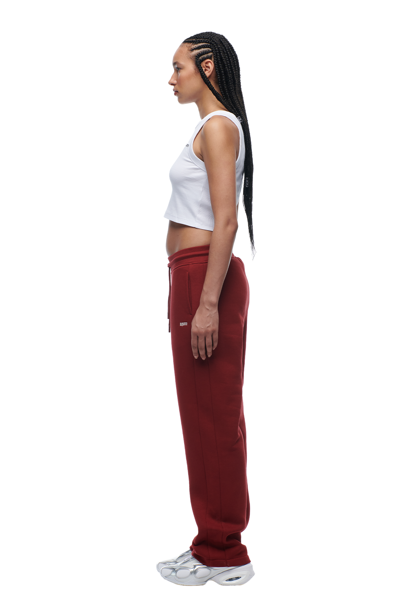 A woman with long braided hair stands in profile wearing the 6PM WMNS OPEN LEG WINE RED sweatpants, a white sleeveless crop top, and white sneakers against a plain background.