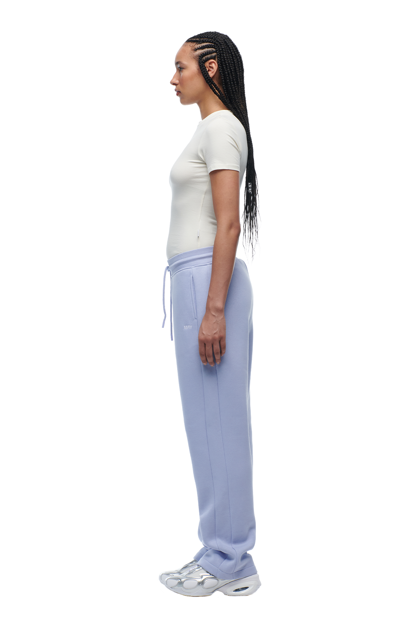 A woman with long braided hair stands in profile against a white background, wearing the 6PM WMNS T-SHIRT OFF-WHITE, light purple sweatpants, and white sneakers.