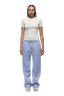 A person with braided hair faces forward, wearing a fitted 6PM WMNS T-SHIRT OFF-WHITE, loose light purple drawstring sweatpants, and white sneakers.