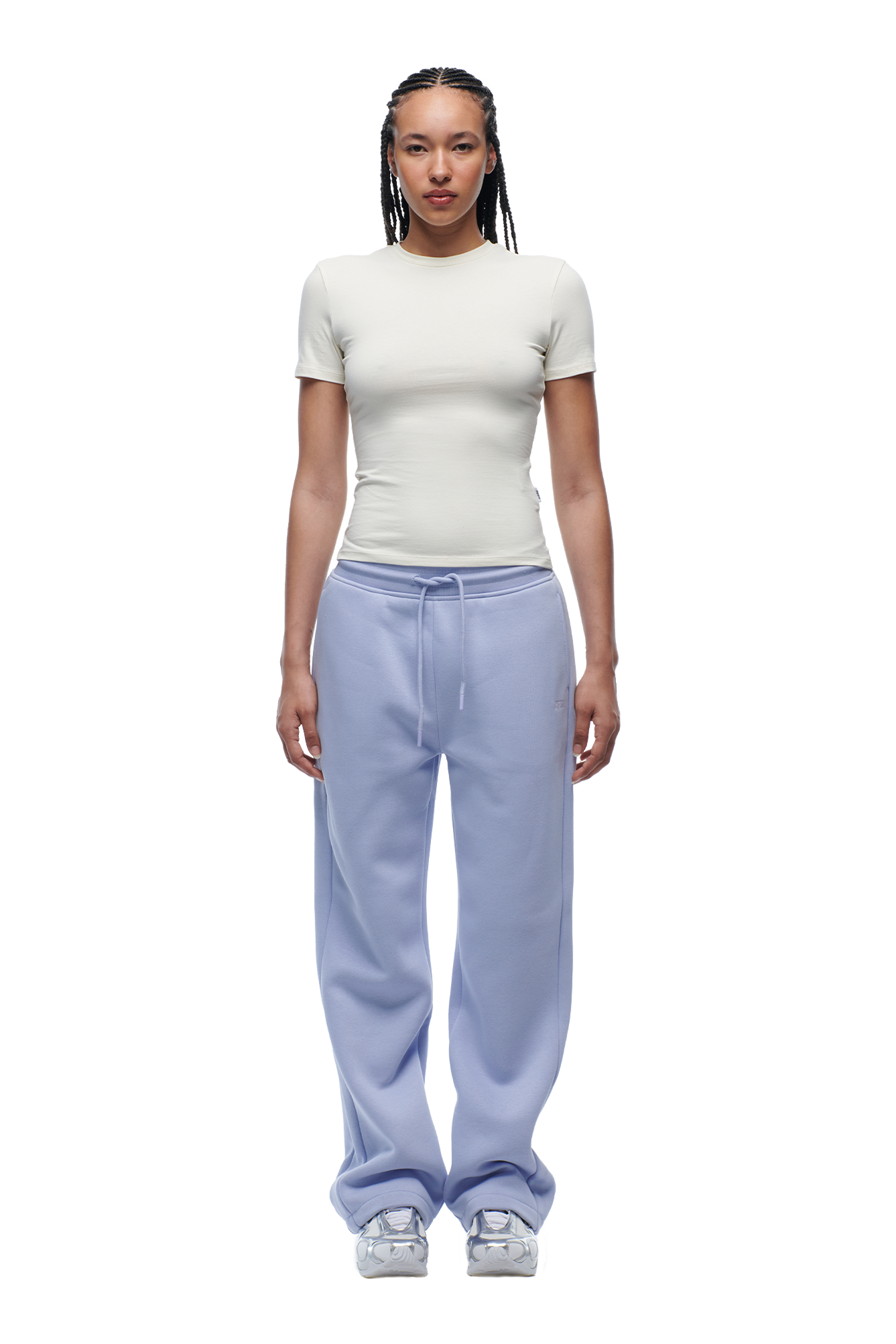 A person with braided hair faces forward, wearing a fitted 6PM WMNS T-SHIRT OFF-WHITE, loose light purple drawstring sweatpants, and white sneakers.