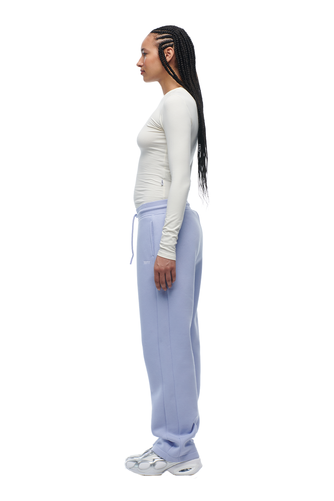 A woman with long braids stands in profile against a white background, wearing the 6PM WMNS LONGSLEEVE OFF-WHITE fitted top, light purple sweatpants with waist ties, and white sneakers.