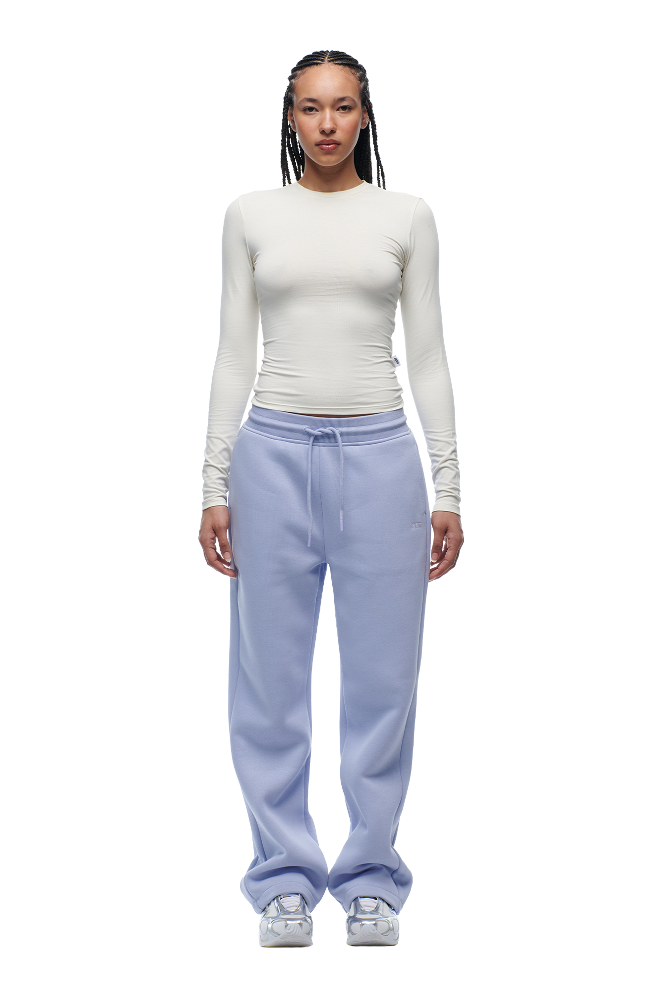A person with braided hair and a neutral expression stands against a white background wearing the 6PM WMNS LONGSLEEVE OFF-WHITE shirt, light blue drawstring sweatpants, and silver sneakers.