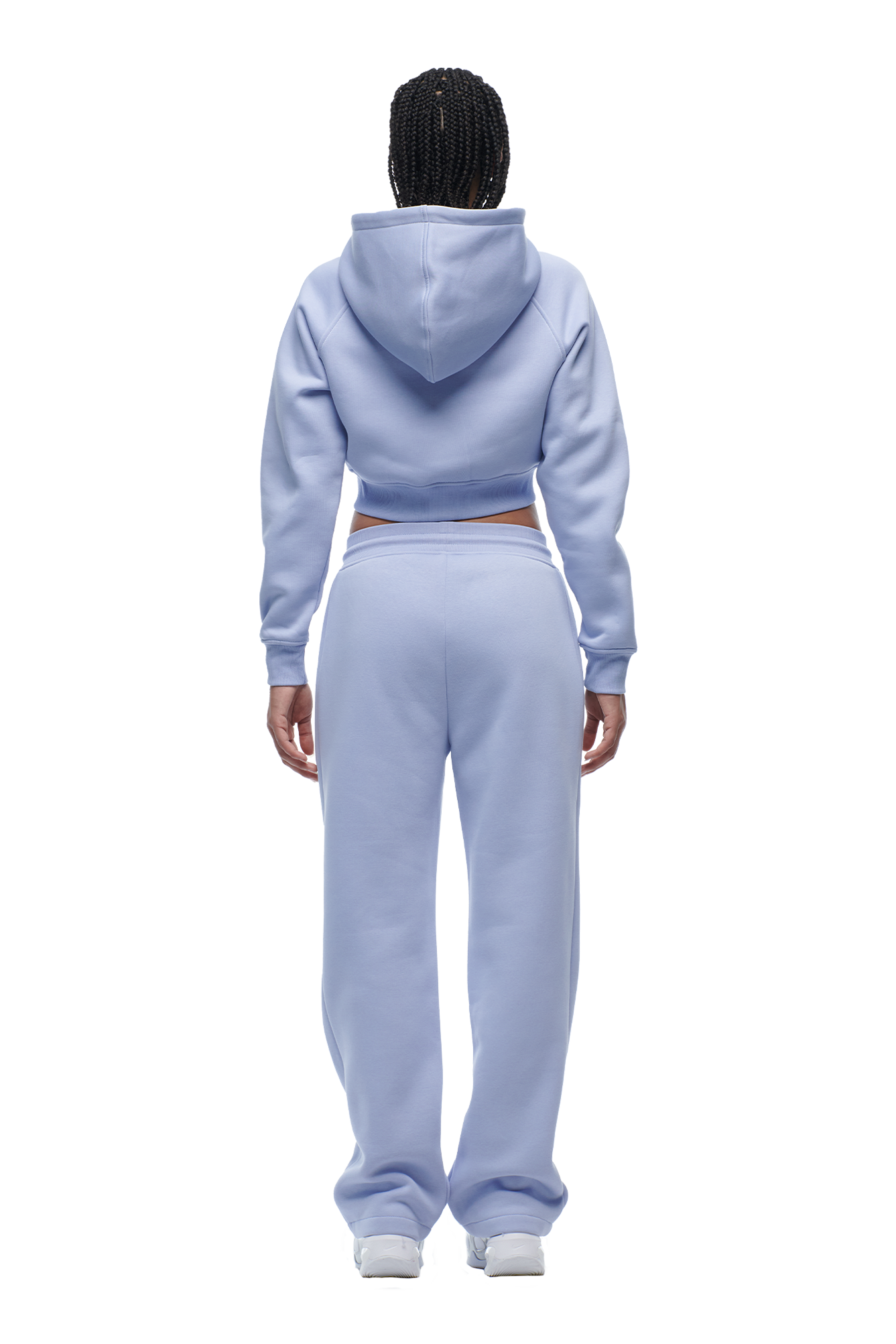 A person with braided hair, facing away, wears the 6PM WMNS Cropped Zip Hoodie in Soft Violet with matching sweatpants and white sneakers. The hoodie’s hood is up and a strip of skin shows at the lower back.