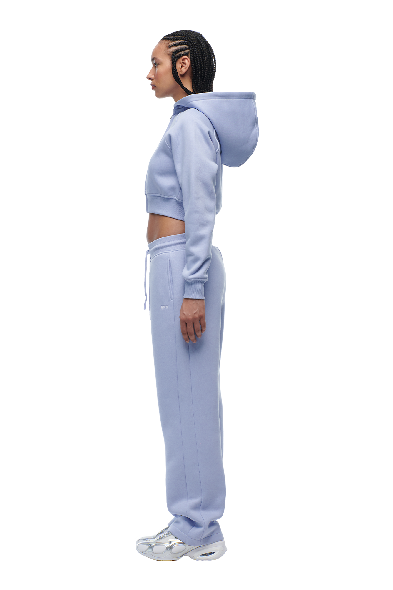 A woman with braided hair stands in profile, wearing the 6PM WMNS CROPPED ZIP HOODIE SOFT VIOLET, matching sweatpants, and white sneakers against a plain white background.