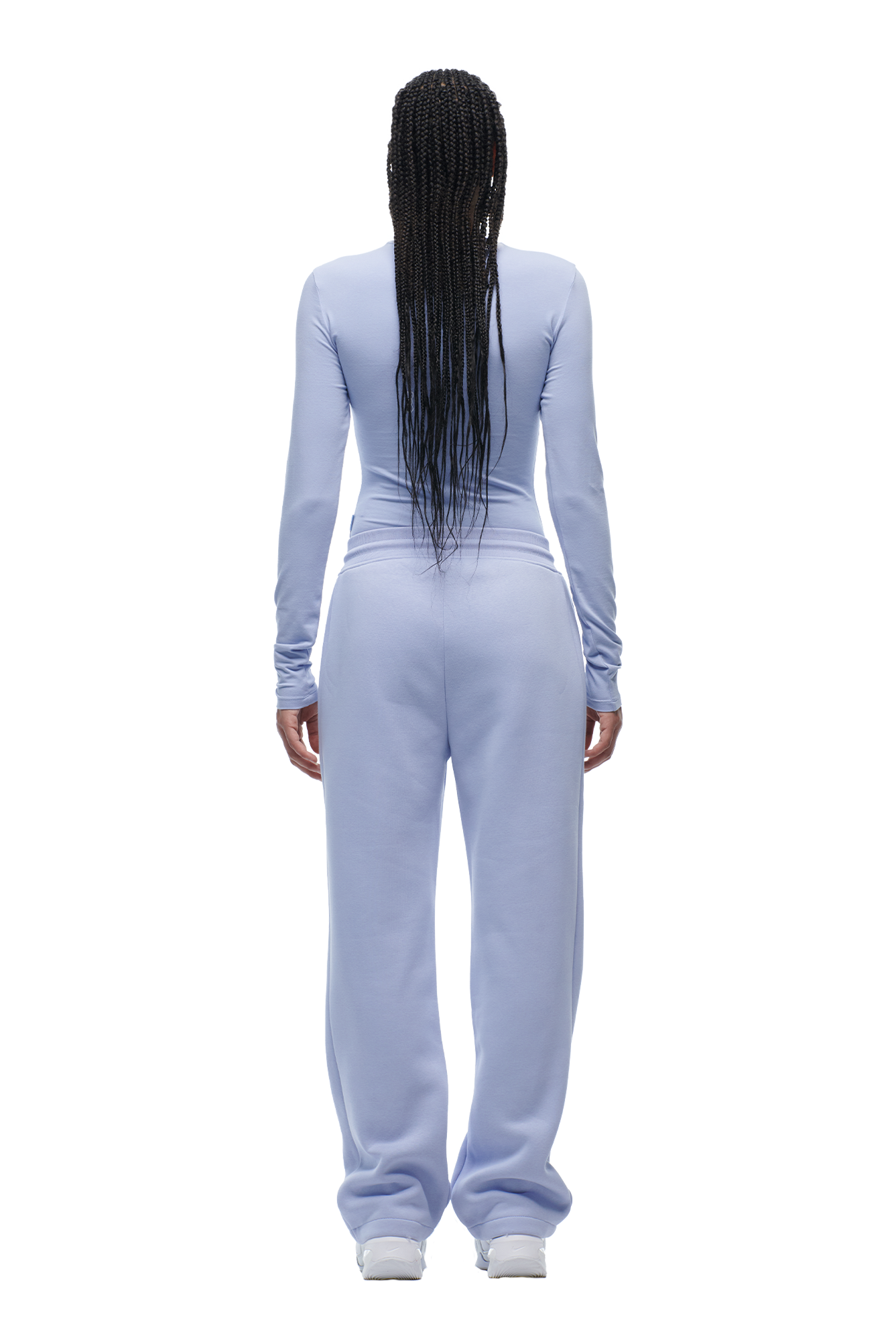 A person with long, braided hair stands with their back to the camera, wearing the 6PM WMNS LONGSLEEVE SOFT VIOLET top and matching sweatpants, paired with white sneakers.