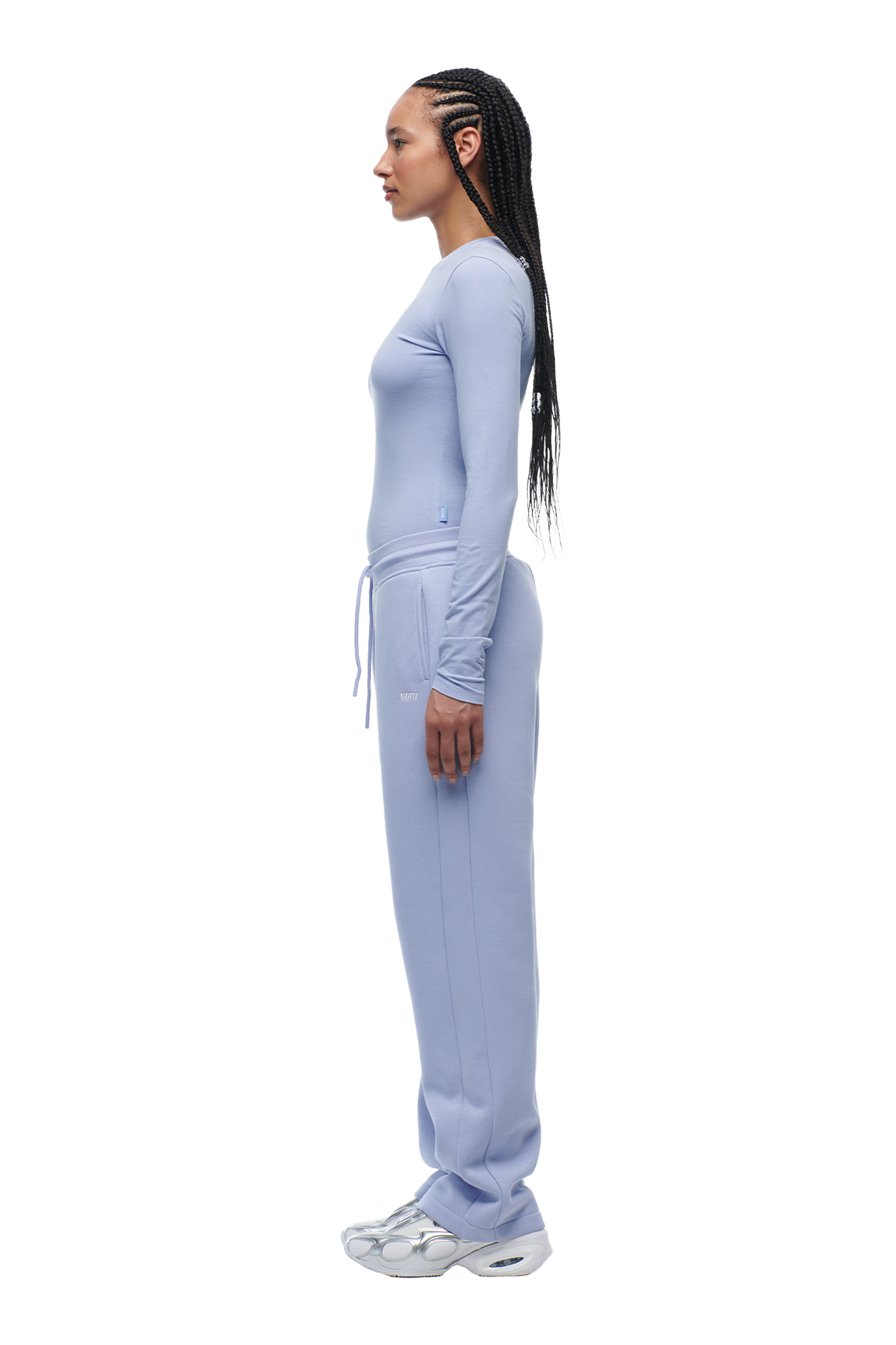 A woman with long braids stands in profile, wearing the 6PM WMNS LONGSLEEVE SOFT VIOLET top with matching sweatpants and white sneakers against a white background.