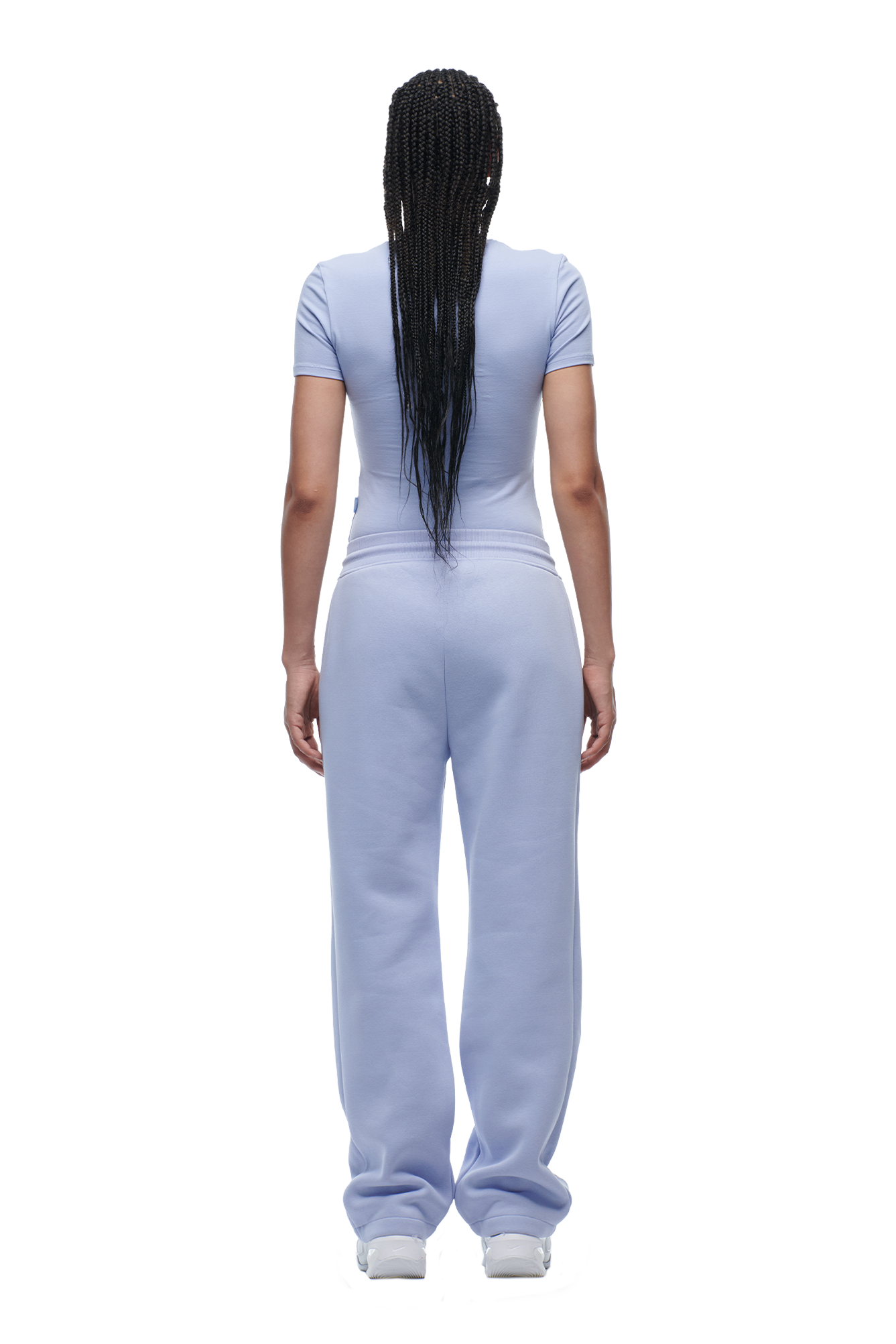 A person with long, braided hair stands facing away, wearing 6PMs WMNS SHORTS SOFT VIOLET and white sneakers.
