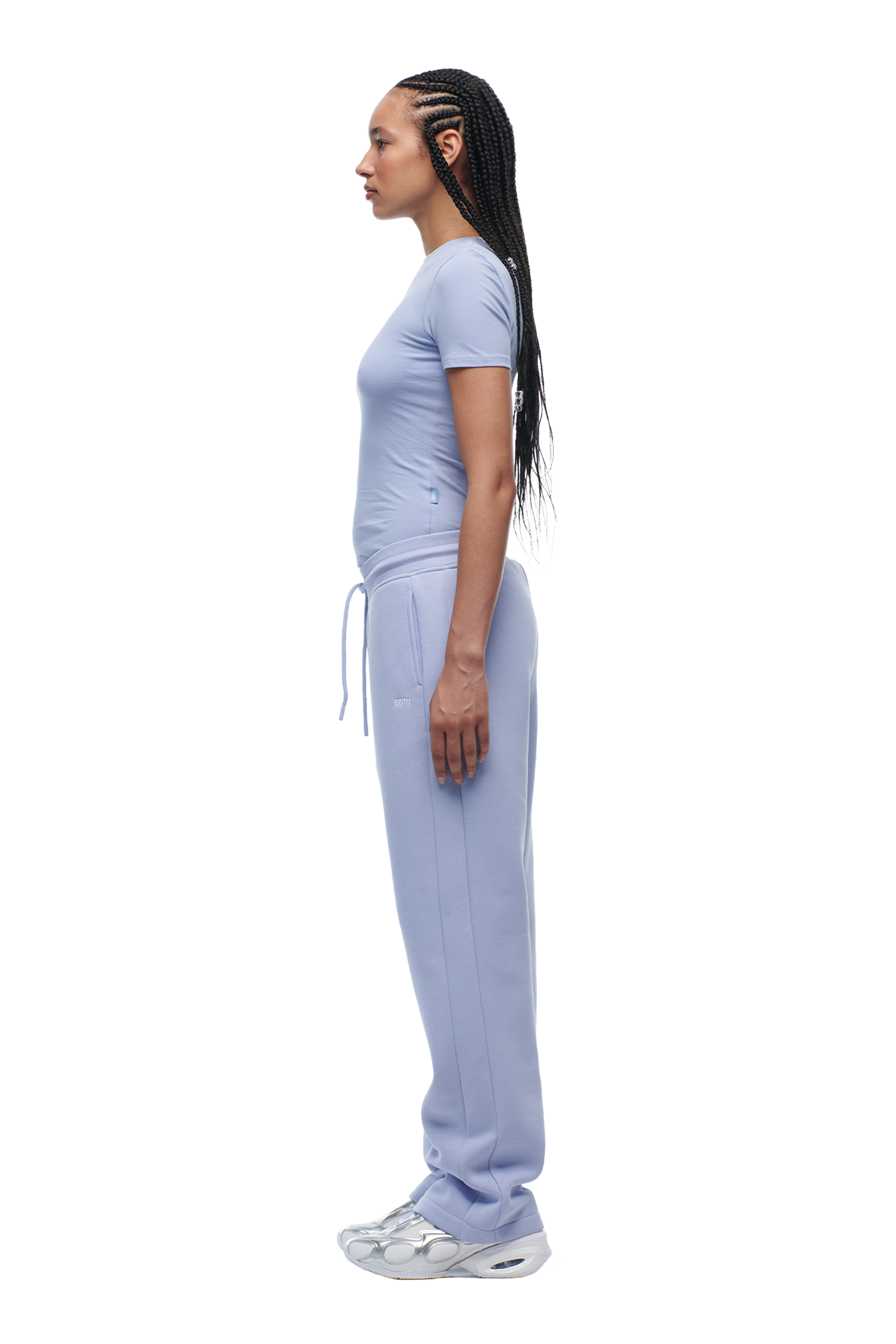 A woman with long braided hair stands in profile facing left against a white background, wearing 6PMs WMNS SHORTS SOFT VIOLET and a fitted matching t-shirt with white sneakers.