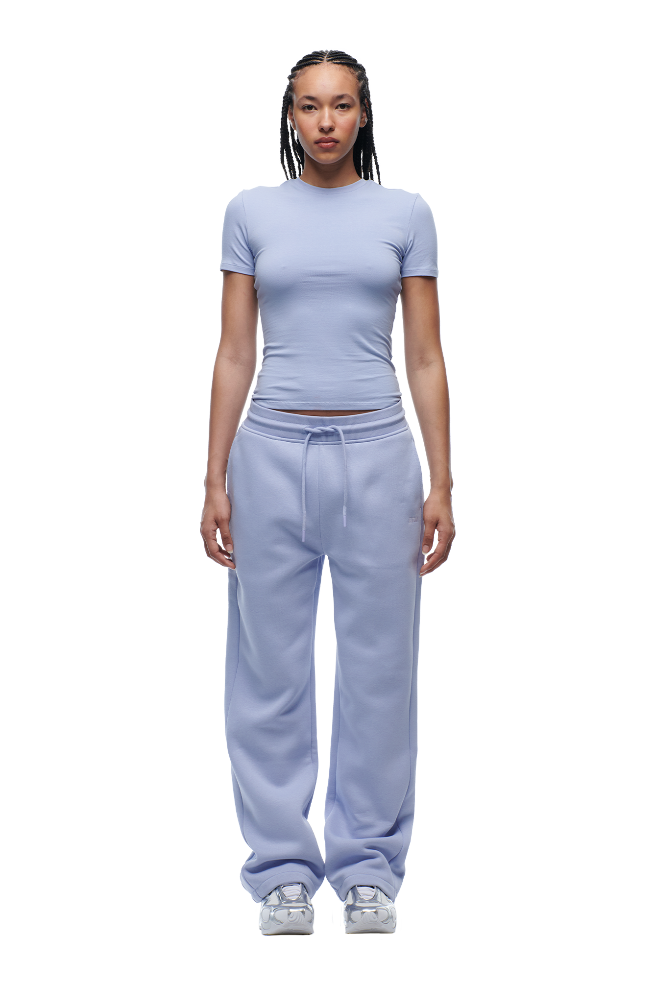 A person stands facing forward, wearing 6PM WMNS SHORTS SOFT VIOLET and a fitted light purple t-shirt with white sneakers. Their hair is braided and they have a neutral expression.
