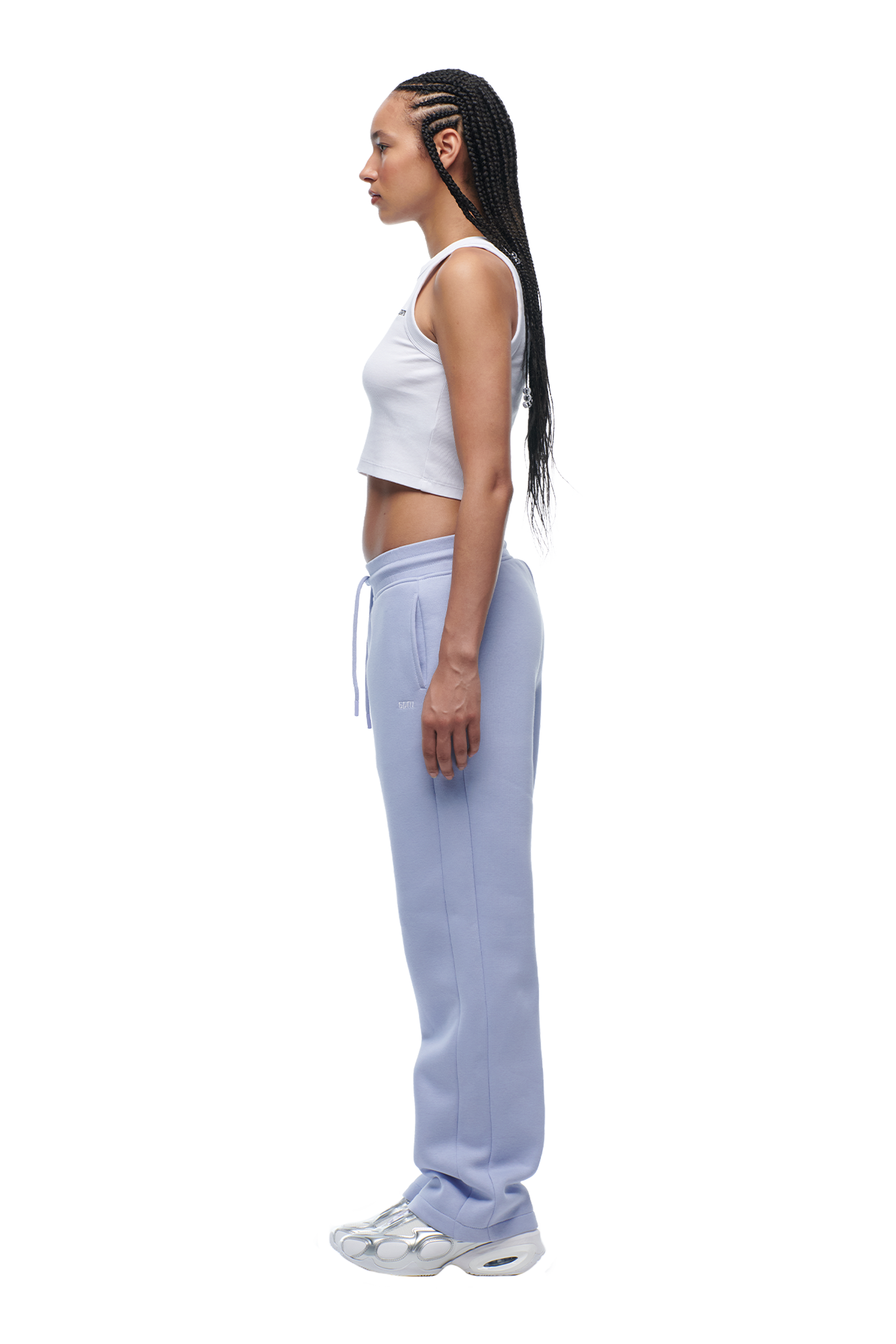 A woman with long braided hair stands in profile against a white background, wearing the 6PM WMNS OPEN LEG SOFT VIOLET sweatpants, a white crop top, and white sneakers.