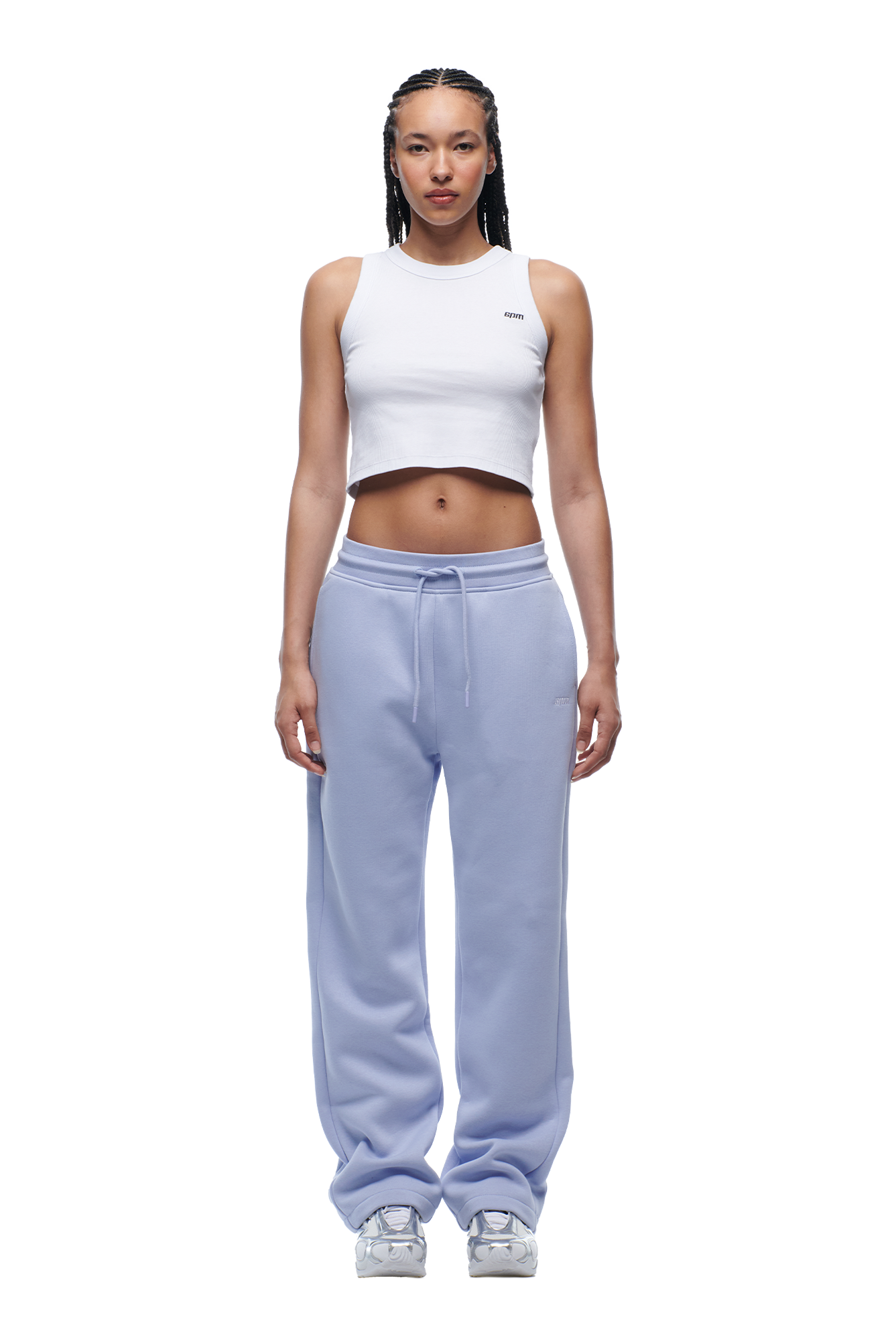 A woman with braided hair and a neutral expression faces forward in a white setting, wearing the 6PM WMNS OPEN LEG SOFT VIOLET pants with a white cropped tank top and white sneakers.