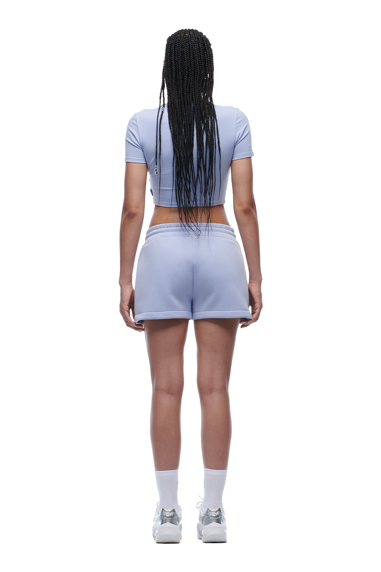 A woman with long braided hair stands facing away, wearing the 6PM WMNS T-SHIRT SOFT VIOLET, matching shorts with white trim, white socks, and sneakers.