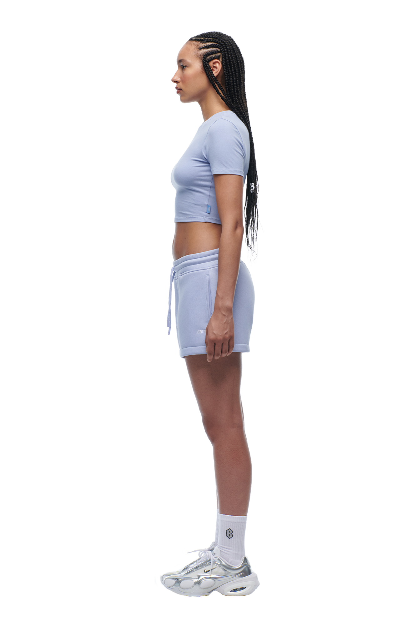 A woman with long braided hair stands in profile against a white background, wearing the 6PM WMNS T-SHIRT SOFT VIOLET crop top, matching shorts, white socks, and silver athletic sneakers. She looks straight ahead.