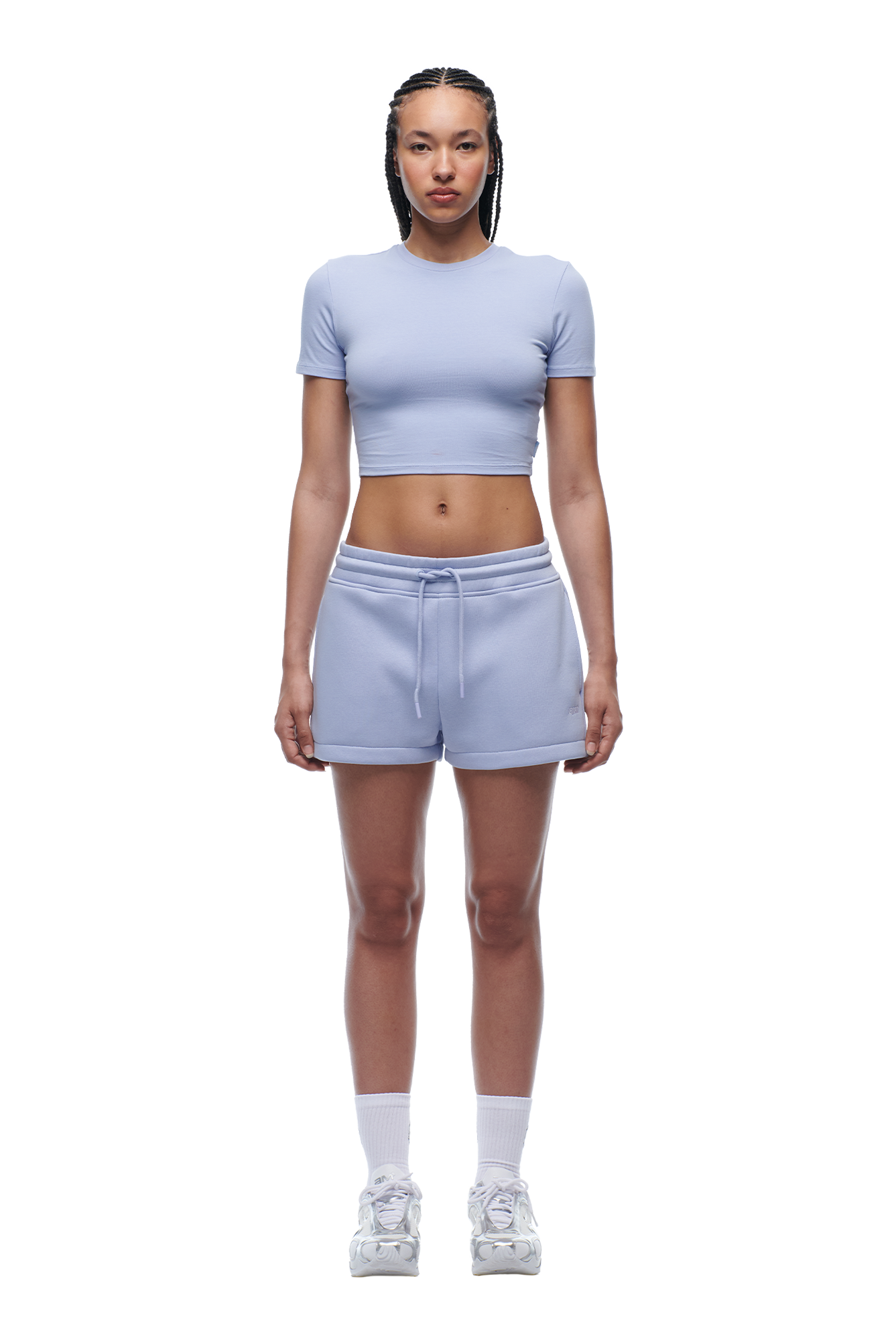 A woman with braided hair and a neutral expression faces forward, wearing the 6PM WMNS T-SHIRT SOFT VIOLET, matching drawstring shorts, white socks, and white sneakers.