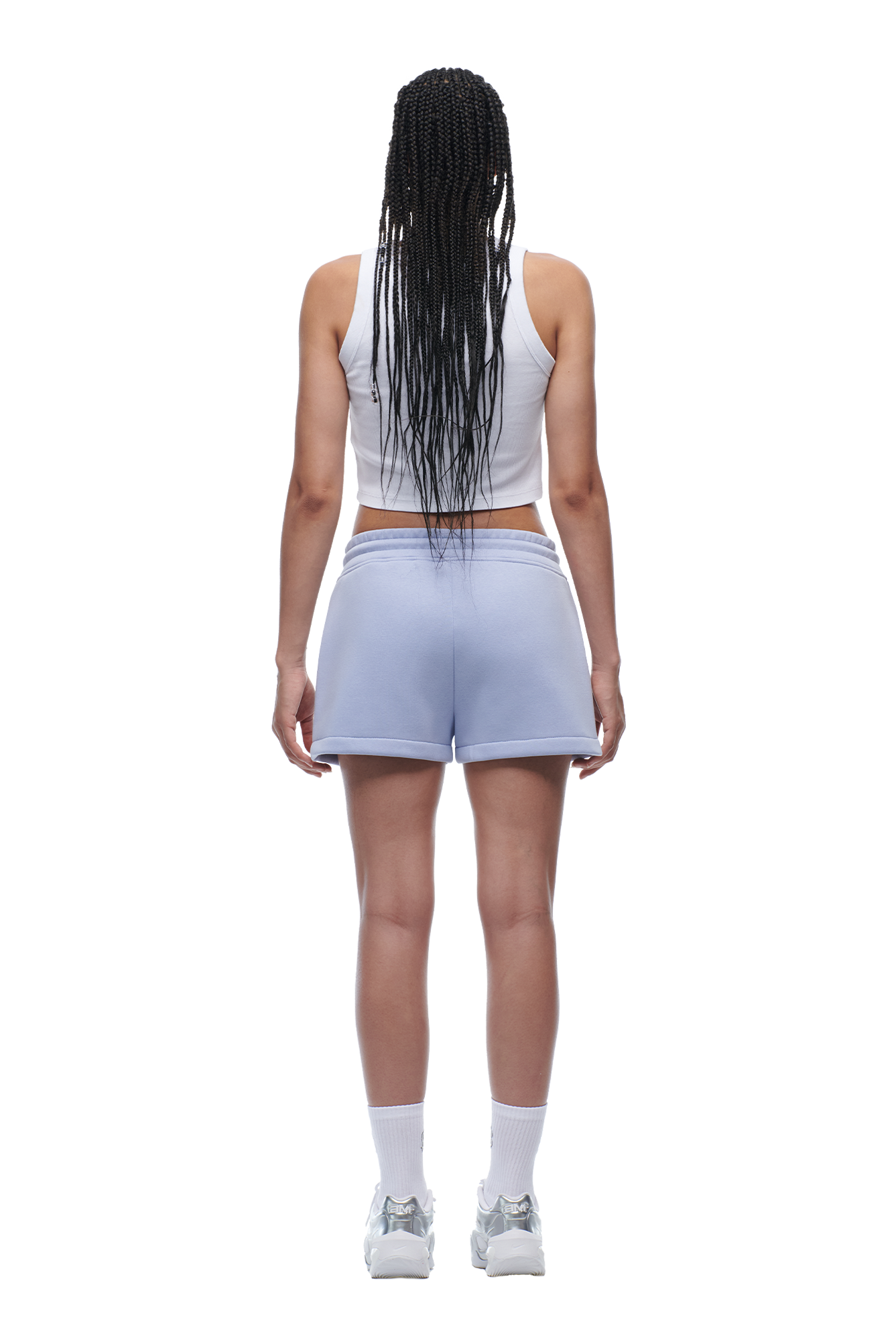 A woman with long braids stands facing away, wearing the 6PM WMNS Cropped T-Shirt in Soft Violet, light blue shorts with white trim, white crew socks, and white sneakers.