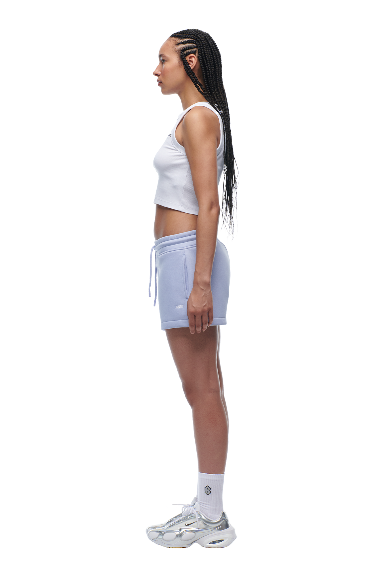 A woman with long braided hair stands in profile against a plain background, wearing the 6PM WMNS CROPPED T-SHIRT SOFT VIOLET, light purple drawstring shorts, white socks, and white sneakers.