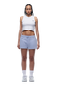 A woman with braided hair and a neutral expression faces forward, wearing the 6PM WMNS Cropped T-Shirt in Soft Violet, light blue drawstring shorts, white crew socks, and white sneakers against a white background.