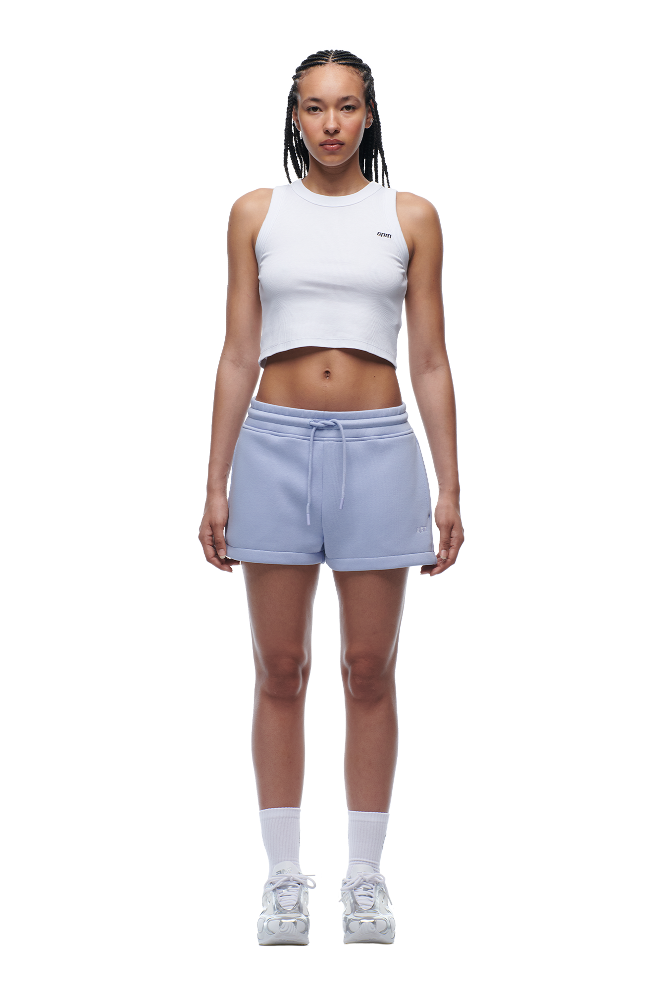 A woman with braided hair and a neutral expression faces forward, wearing the 6PM WMNS Cropped T-Shirt in Soft Violet, light blue drawstring shorts, white crew socks, and white sneakers against a white background.