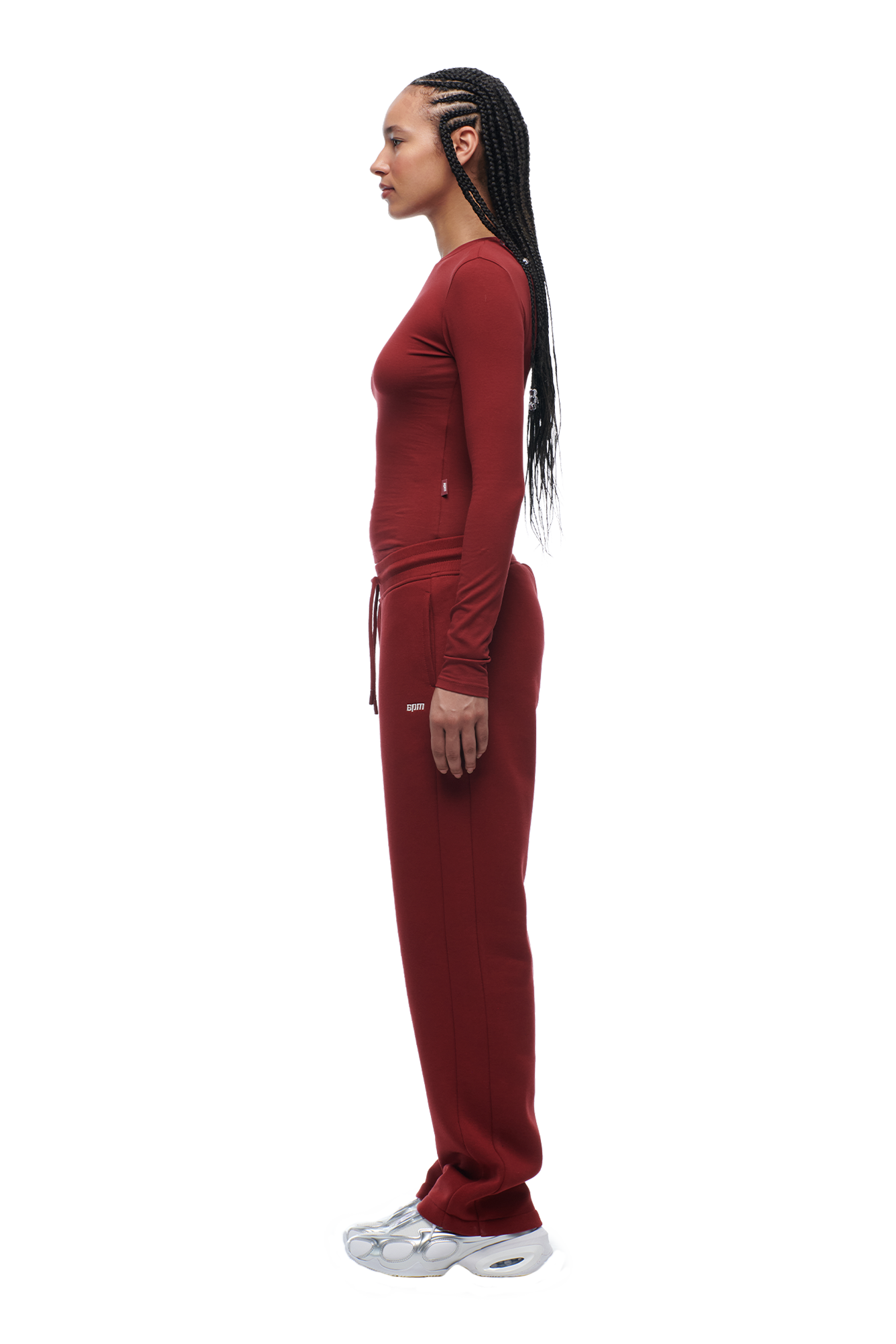 A woman with long braids stands in profile, wearing the 6PM WMNS LONGSLEEVE WINE RED top and matching pants with white sneakers against a white background.