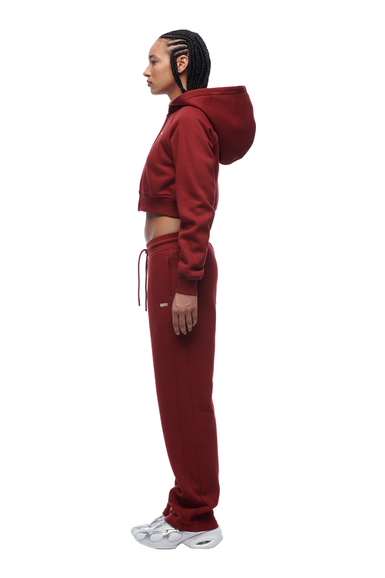 A woman with cornrows stands in profile, arms relaxed, wearing the 6PM WMNS Cropped Zip Hoodie Wine Red with matching sweatpants and white sneakers.
