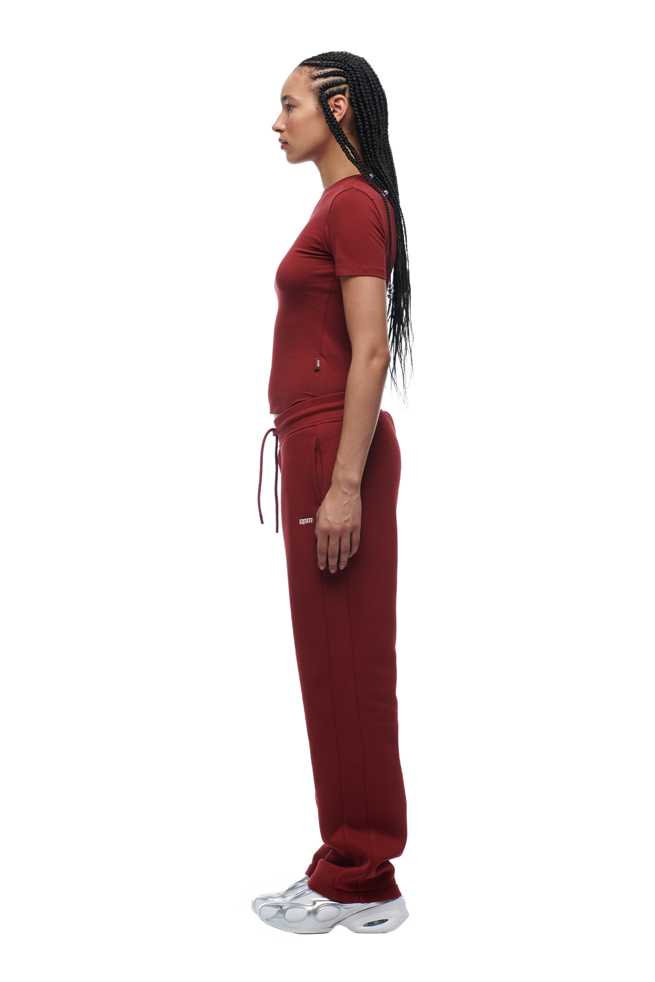 A woman with long braids stands in profile against a white background, wearing the 6PM WMNS T-SHIRT WINE RED, matching maroon sweatpants, and white sneakers.