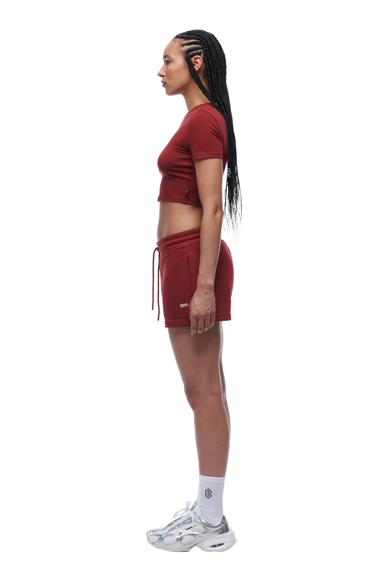 A woman with long braided hair stands in profile against a white background, wearing the 6PM WMNS CROPPED T-SHIRT WINE RED, matching shorts, white socks, and silver athletic shoes.