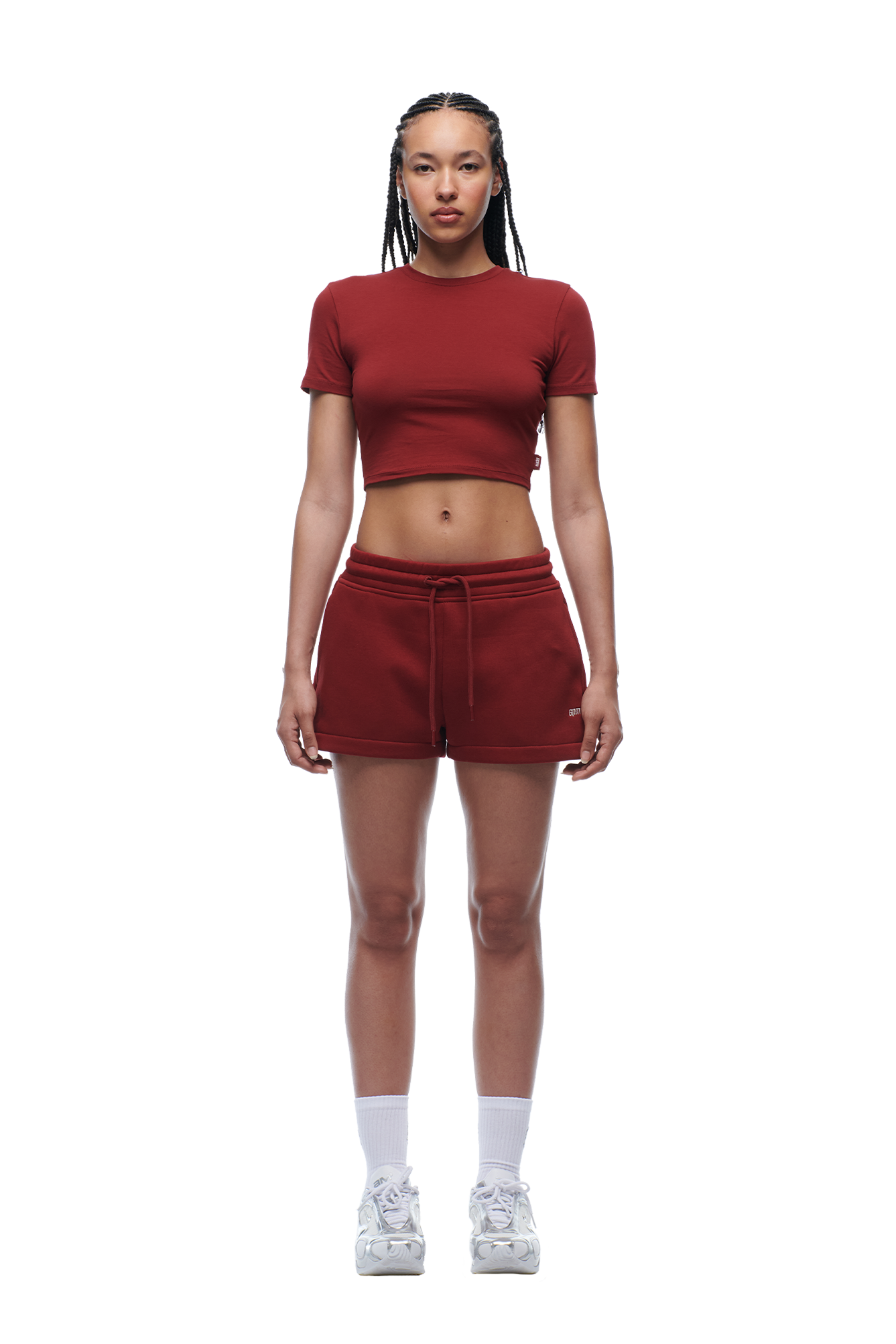 A woman with braided hair wears the 6PM WMNS CROPPED T-SHIRT WINE RED, maroon shorts, white socks, and white sneakers, standing facing forward with a neutral expression against a white background.