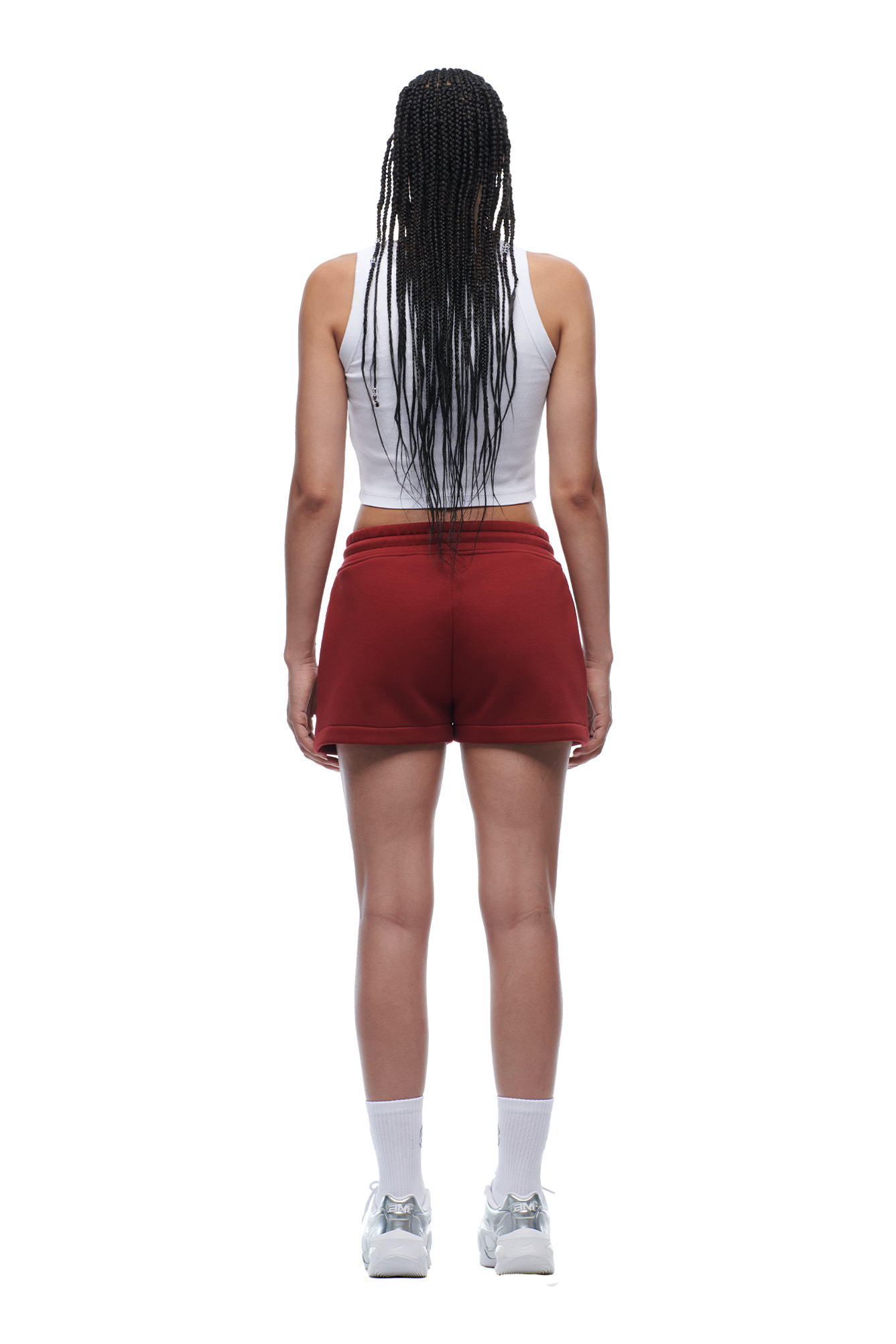 A person with long braided hair, wearing a white tank top, 6PM WMNS SHORTS WINE RED, white socks, and silver sneakers is standing and facing away from the camera.