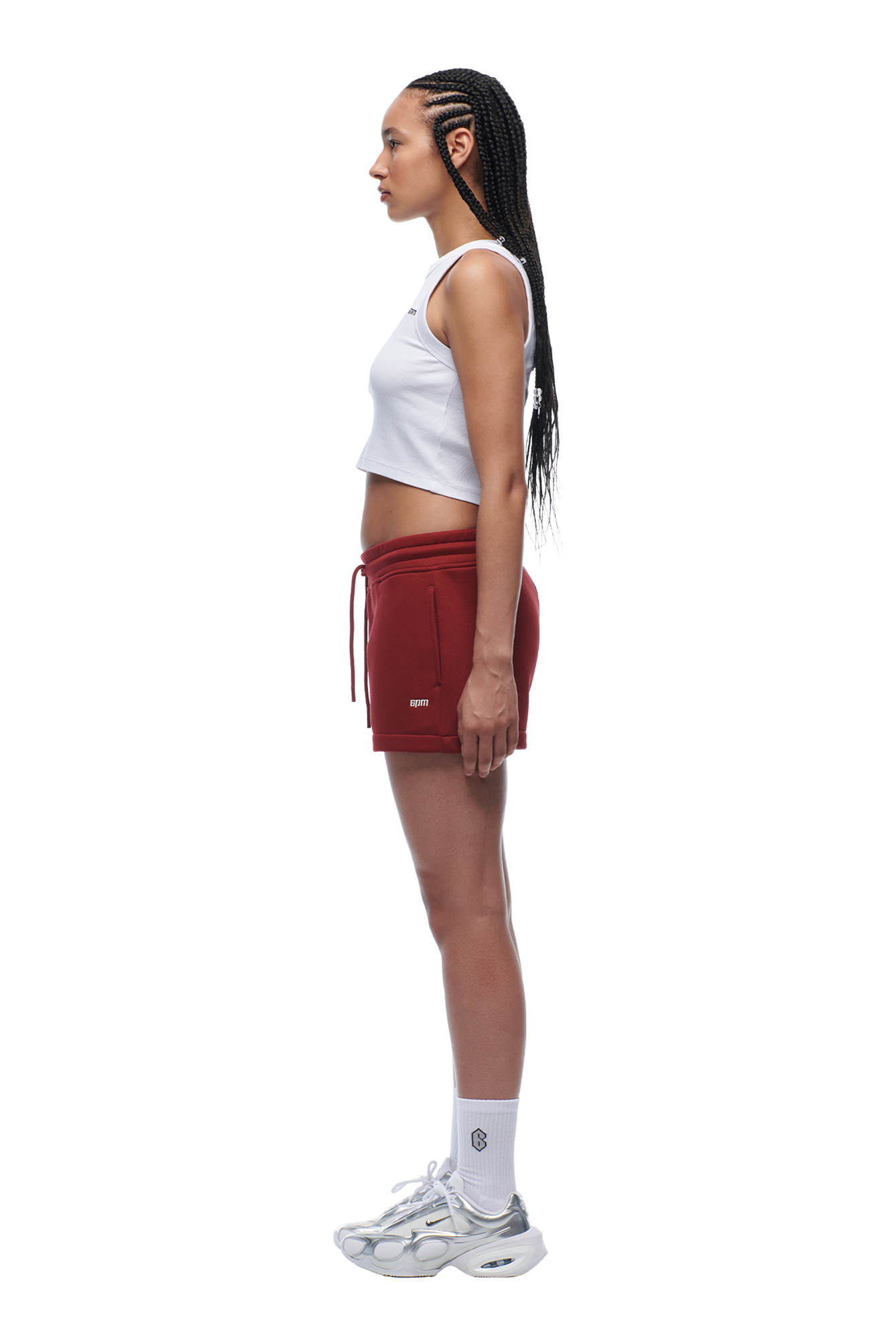 A woman with long braided hair stands in profile wearing a white crop top, 6PM WMNS SHORTS WINE RED, white socks, and white sneakers against a plain white background.