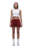 A woman with braided hair and a neutral expression faces forward, wearing a white cropped tank top, white sneakers and crew socks, and 6PM WMNS SHORTS WINE RED.