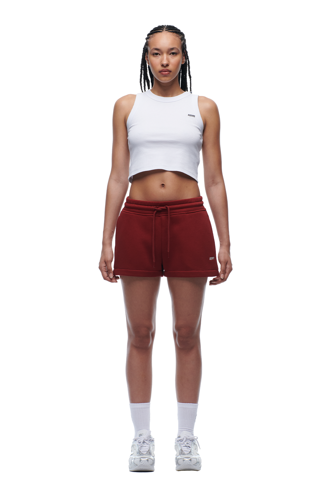 A woman with braided hair and a neutral expression faces forward, wearing a white cropped tank top, white sneakers and crew socks, and 6PM WMNS SHORTS WINE RED.