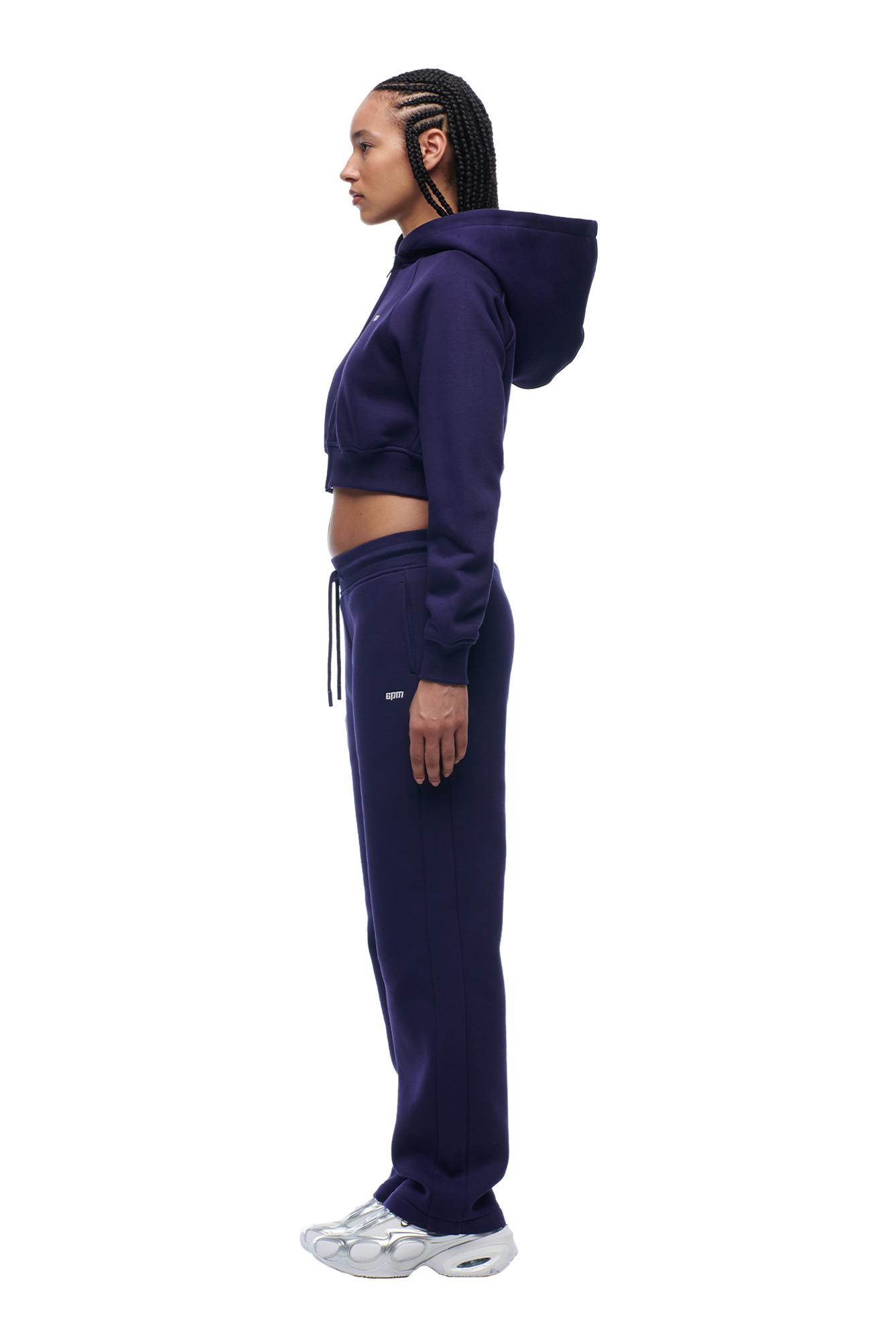 A woman with braided hair stands in profile on a white background, wearing the 6PM WMNS Cropped Zip Hoodie in Orbit Blue, paired with matching sweatpants and white sneakers.