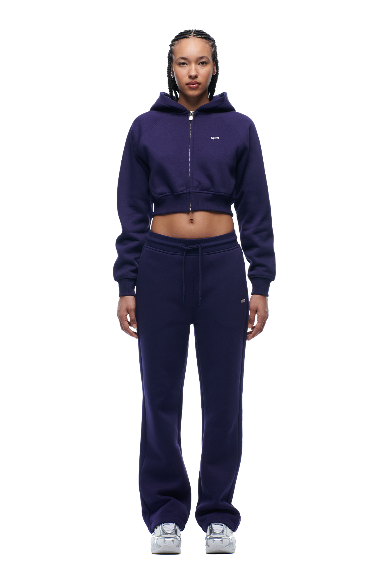 A woman with braided hair and a neutral expression faces forward, wearing the 6PM WMNS Cropped Zip Hoodie in Orbit Blue with matching drawstring sweatpants and white sneakers.
