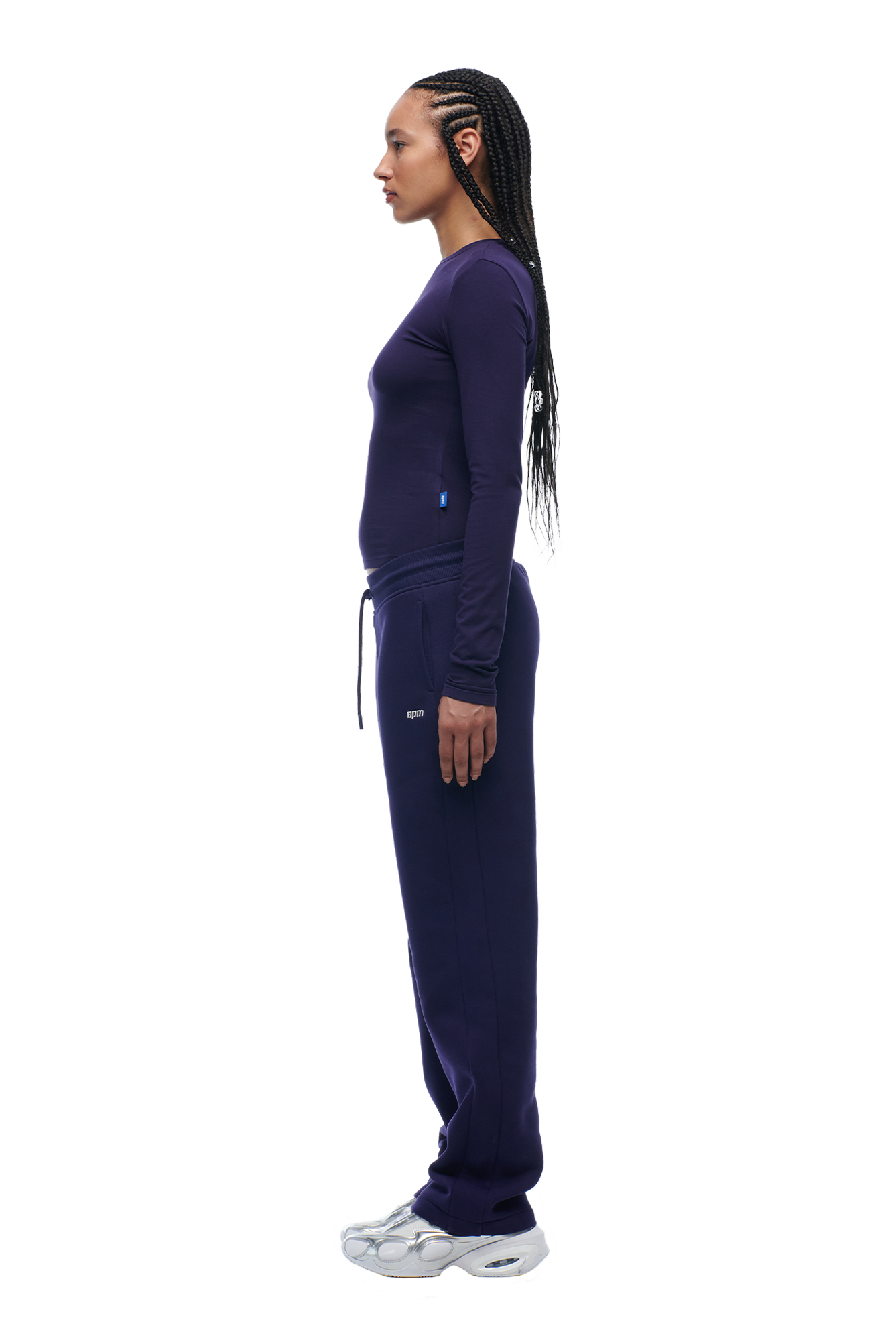 A person with long braided hair wears the 6PM WMNS LONGSLEEVE ORBIT BLUE top, matching navy sweatpants, and white sneakers, shown in profile against a plain white background.