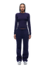 A woman with braided hair and a neutral expression faces forward, wearing the 6PM WMNS LONGSLEEVE ORBIT BLUE top, matching drawstring sweatpants, and white sneakers.