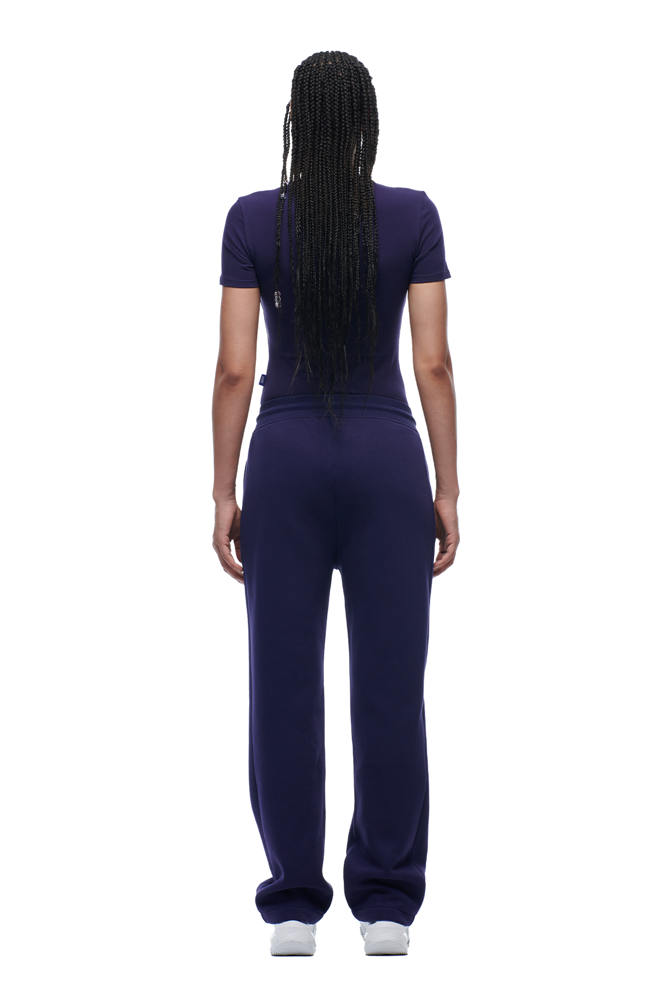 A person with long braided hair, wearing the 6PM WMNS T-SHIRT ORBIT BLUE and loose purple sweatpants, stands facing away from the camera against a plain white background.
