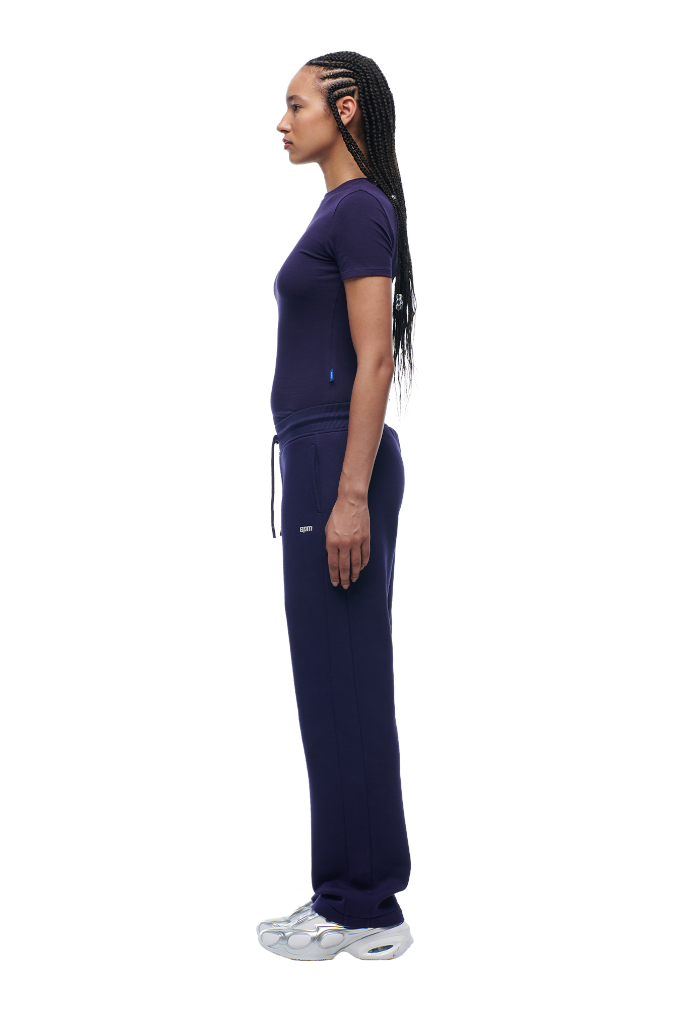 A woman with long braided hair stands in profile against a white background, wearing the 6PM WMNS T-SHIRT ORBIT BLUE, matching sweatpants, and white sneakers.