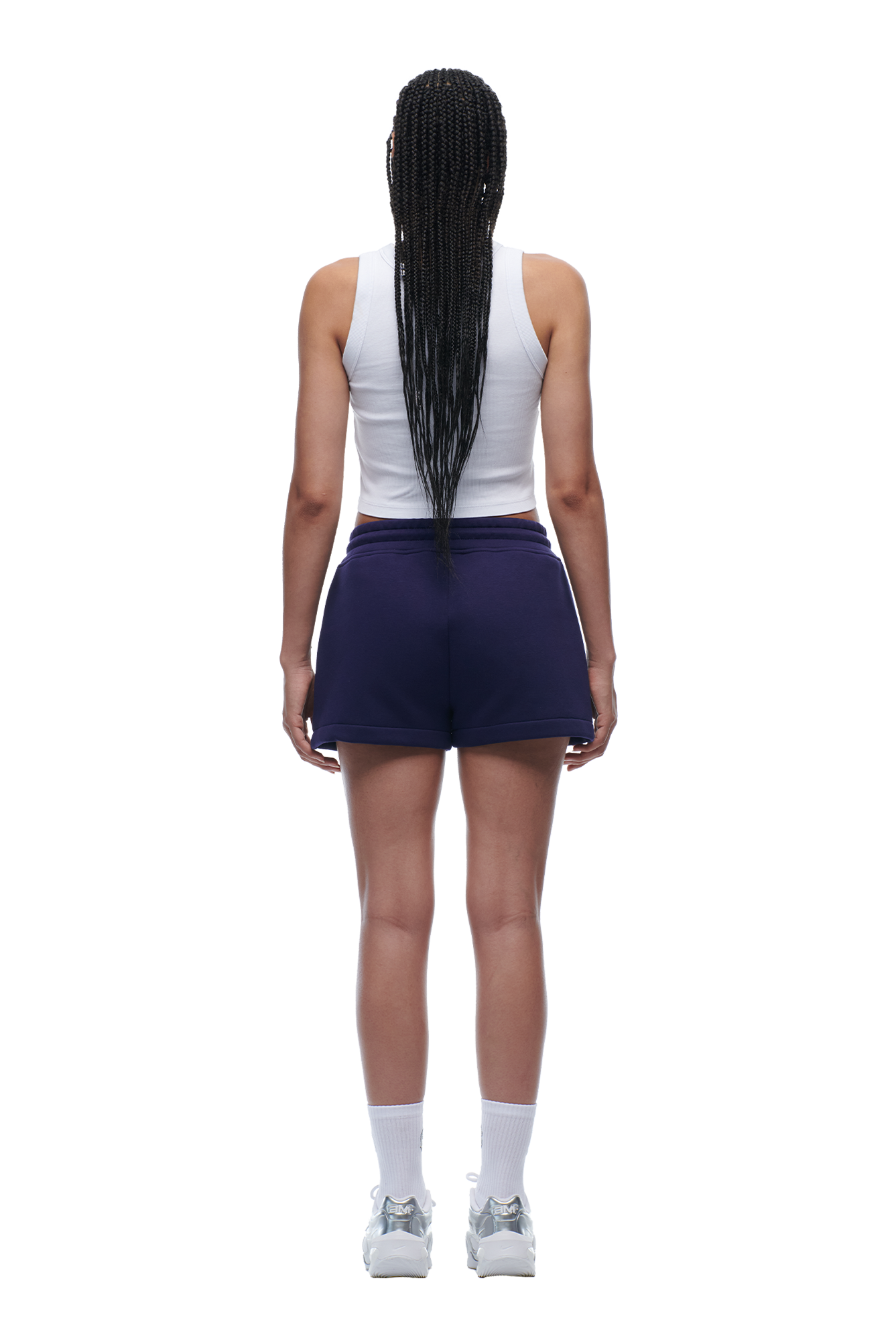 A woman with long braided hair faces away, wearing a white tank top, 6PM WMNS SHORTS ORBIT BLUE, white socks, and sneakers against a white background.