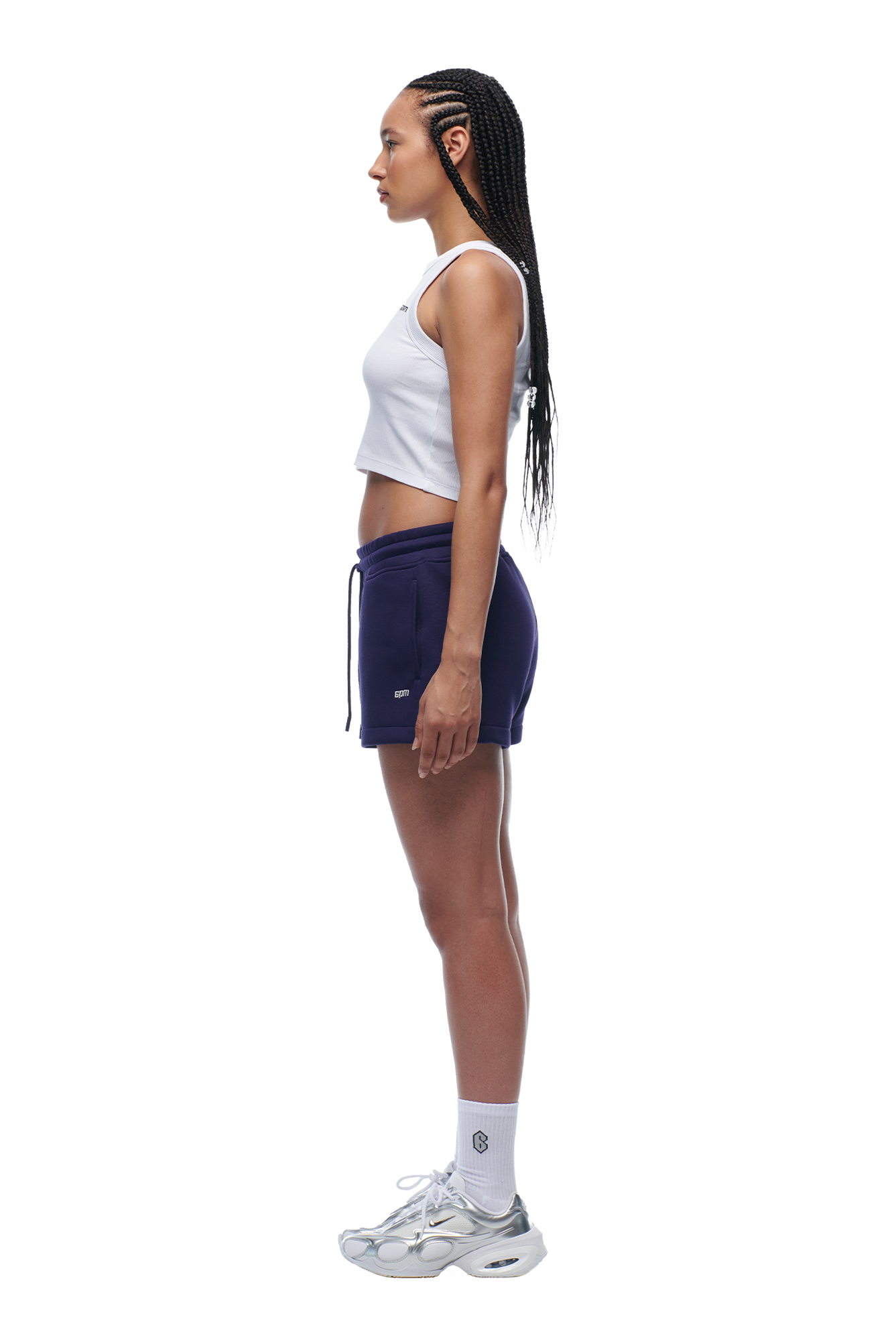 A woman with long braids stands in profile against a plain white background, wearing a white tank top, 6PM WMNS SHORTS ORBIT BLUE, white socks, and white sneakers.