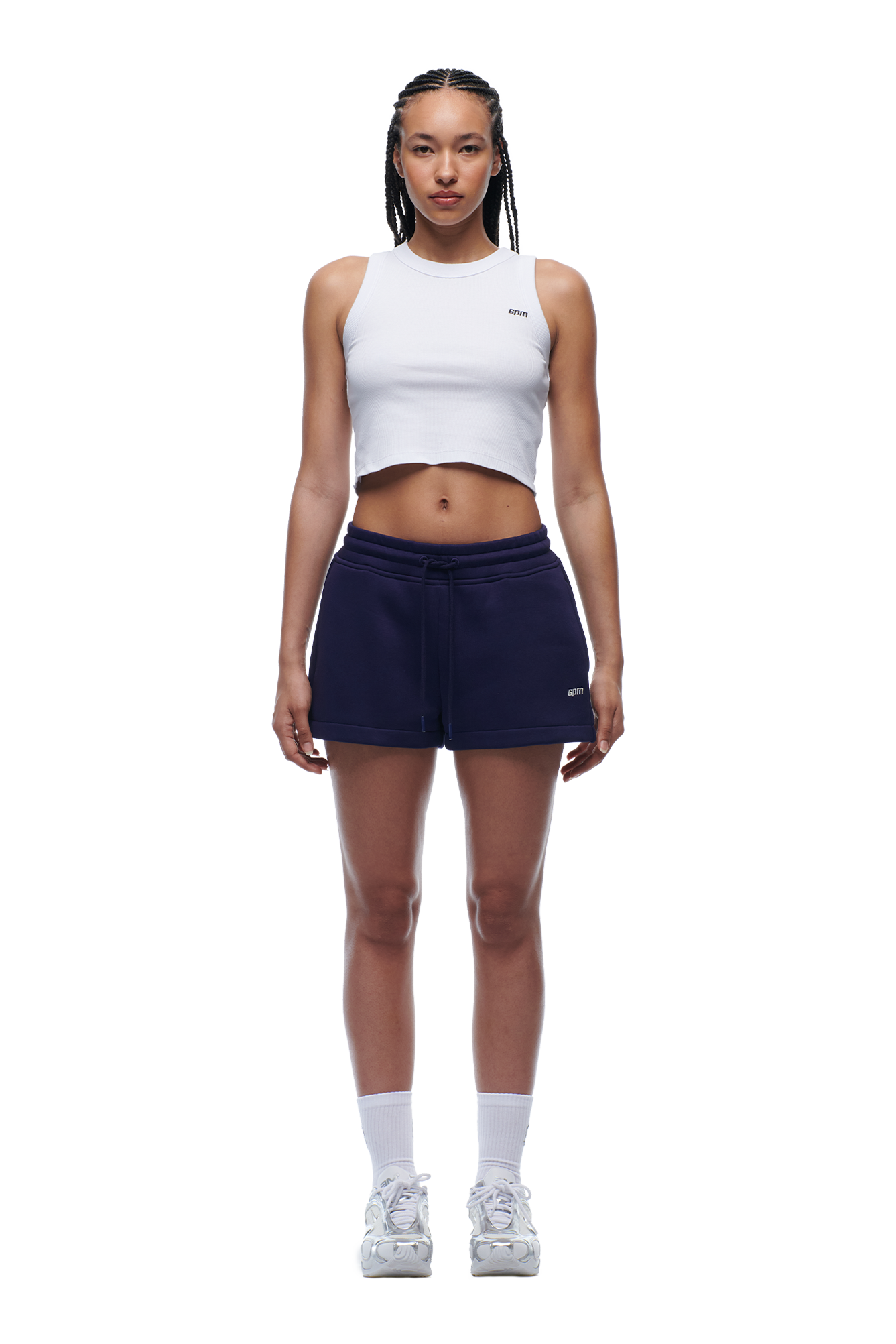 A woman with braided hair and a neutral expression stands facing forward in a white cropped tank, 6PM WMNS SHORTS ORBIT BLUE, white socks, and sneakers against a plain white background.