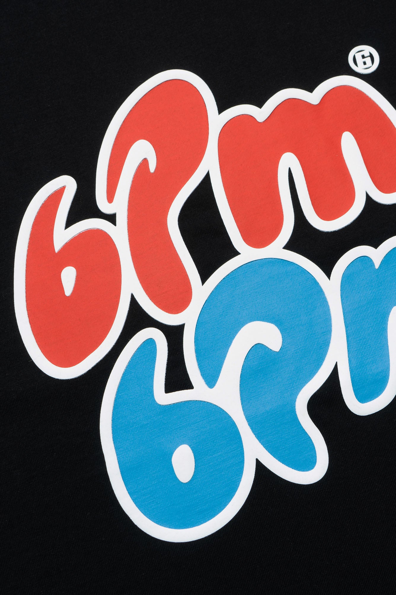 A close-up of bold, stylized “6PM 6PM” text in red and blue bubble letters with white outlines on a black ICE CREAM T-SHIRT by 6PM. A small circled B symbol is in the upper right corner.