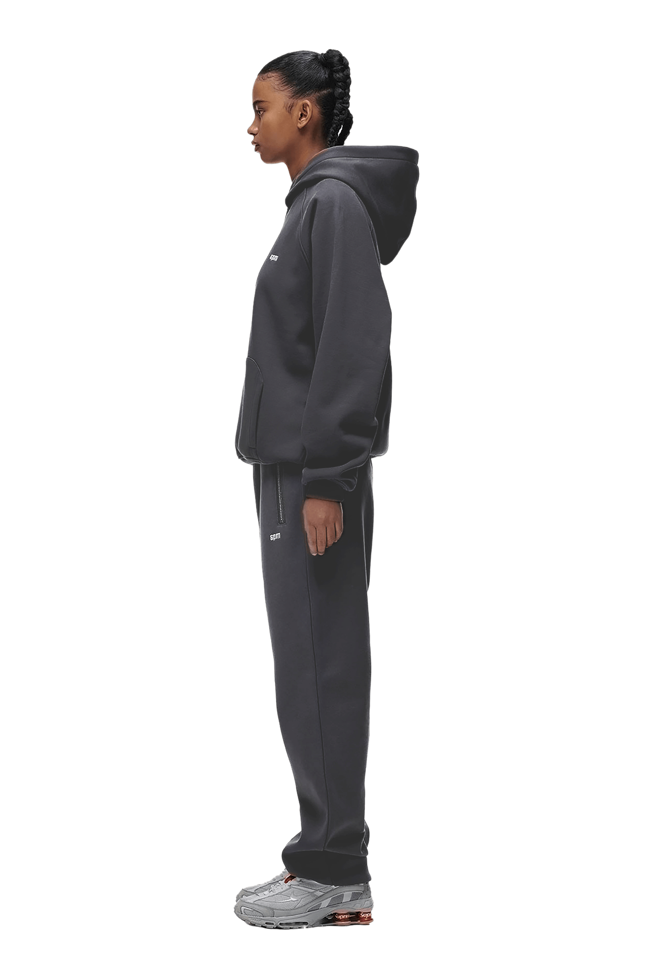 A woman with her hair in a braid stands in profile against a solid green background, wearing the 6PM HOODIE ANTHRACITE with raglan sleeves, a kangaroo pocket, matching sweatpants, and gray sneakers—ideal for everyday wear.