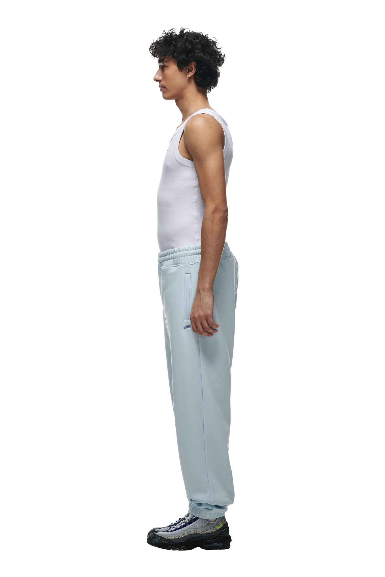 A man with curly hair stands in profile against a white background, wearing a white tank top, black sneakers, and the 6PM CUFF JOGGER ICE BLUE sweatpants with cuffed leg openings.