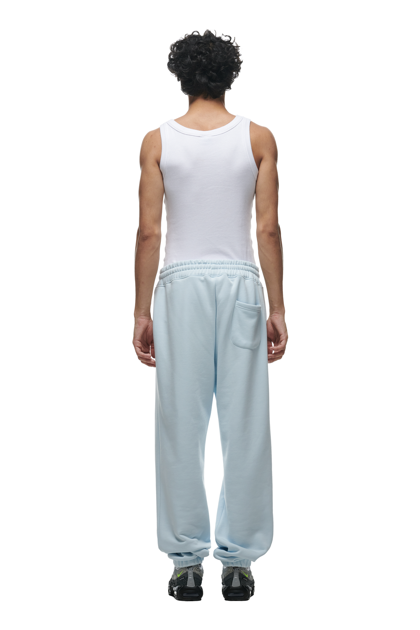 A person with short curly hair faces away, wearing a white sleeveless top and 6PM’s CUFF JOGGER ICE BLUE sweatpants featuring a back pocket and cuffed leg opening, paired with dark sneakers. The background is plain white.