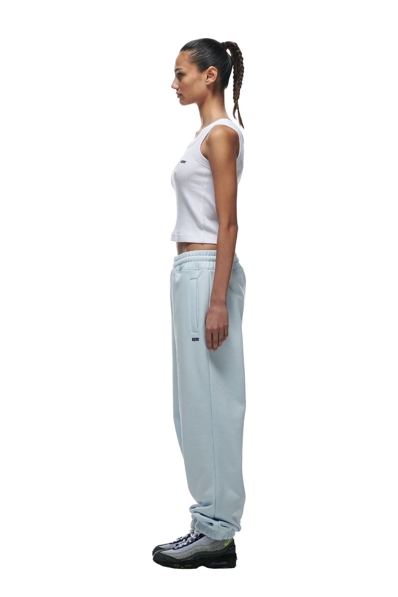 A woman stands in profile wearing a white sleeveless top, 6PM CUFF JOGGER ICE BLUE sweatpants with cuffed legs, and gray sneakers. Her hair is tied back in a ponytail and her arms rest at her sides.